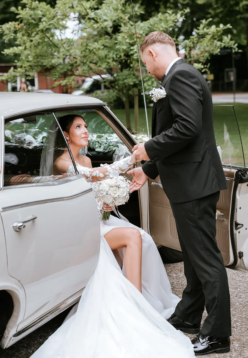 Der Bräutigam reicht der Braut am geschmückten Wagen die Hand, sie steigt aus und hält den Strauß; reportagefotografie hochzeit und natürliche hochzeitsfotografie fangen den Moment ein
