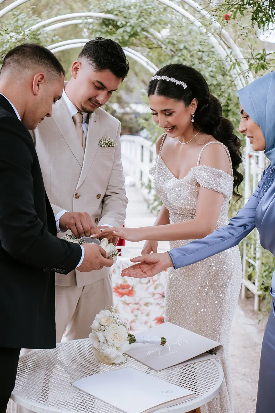Braut und Bräutigam stecken sich die Ringe an, Familie hilft am kleinen Tisch unter einem Laubengang; reportagefotografie hochzeit und fotograf für sommerhochzeit