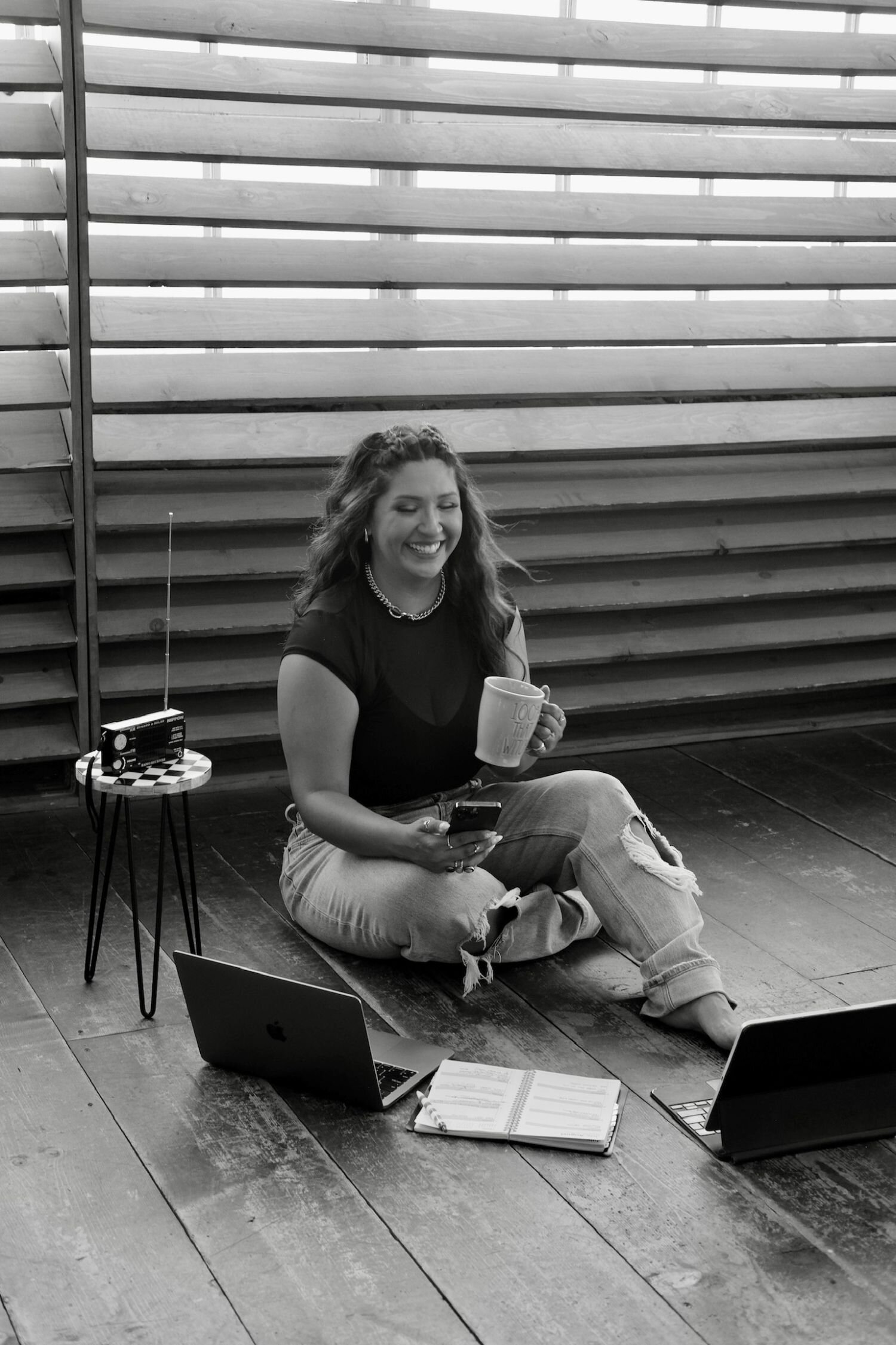 Joyce sitting in the floor holding a mug. She’s wearing a silver choker and hoop earrings, a sheer black top, and ripped jeans.