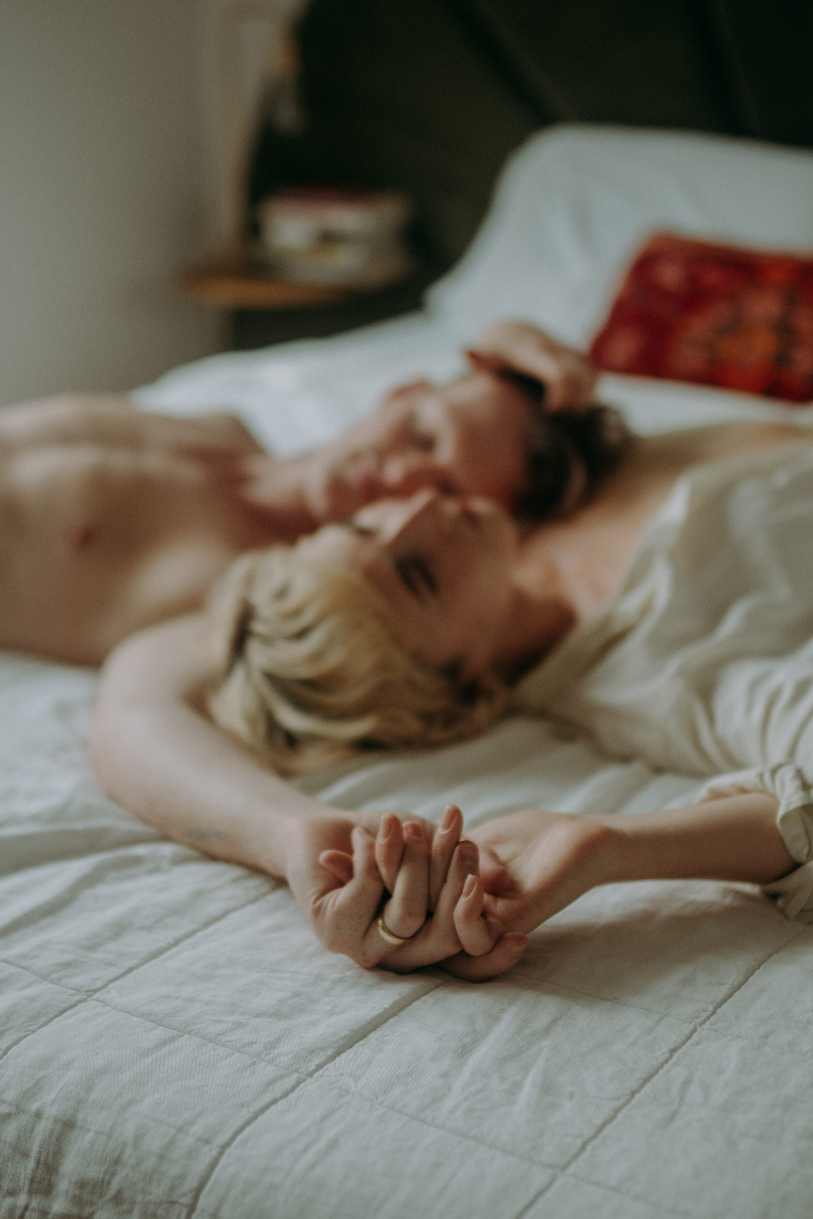 Couple lying on a bed, holding hands, with their faces soft and intimate in the background.