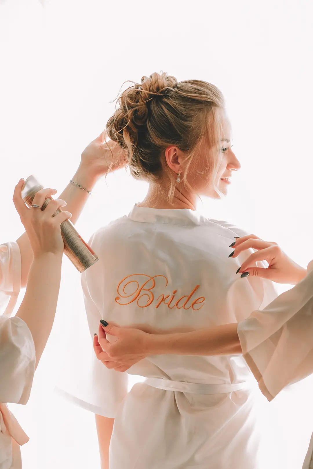 Braut im Morgenmantel am Fenster; Hände richten den Stoff, Haarspray fixiert den Dutt – getting ready fotografie und natürliche hochzeitsfotografie in Hamburg