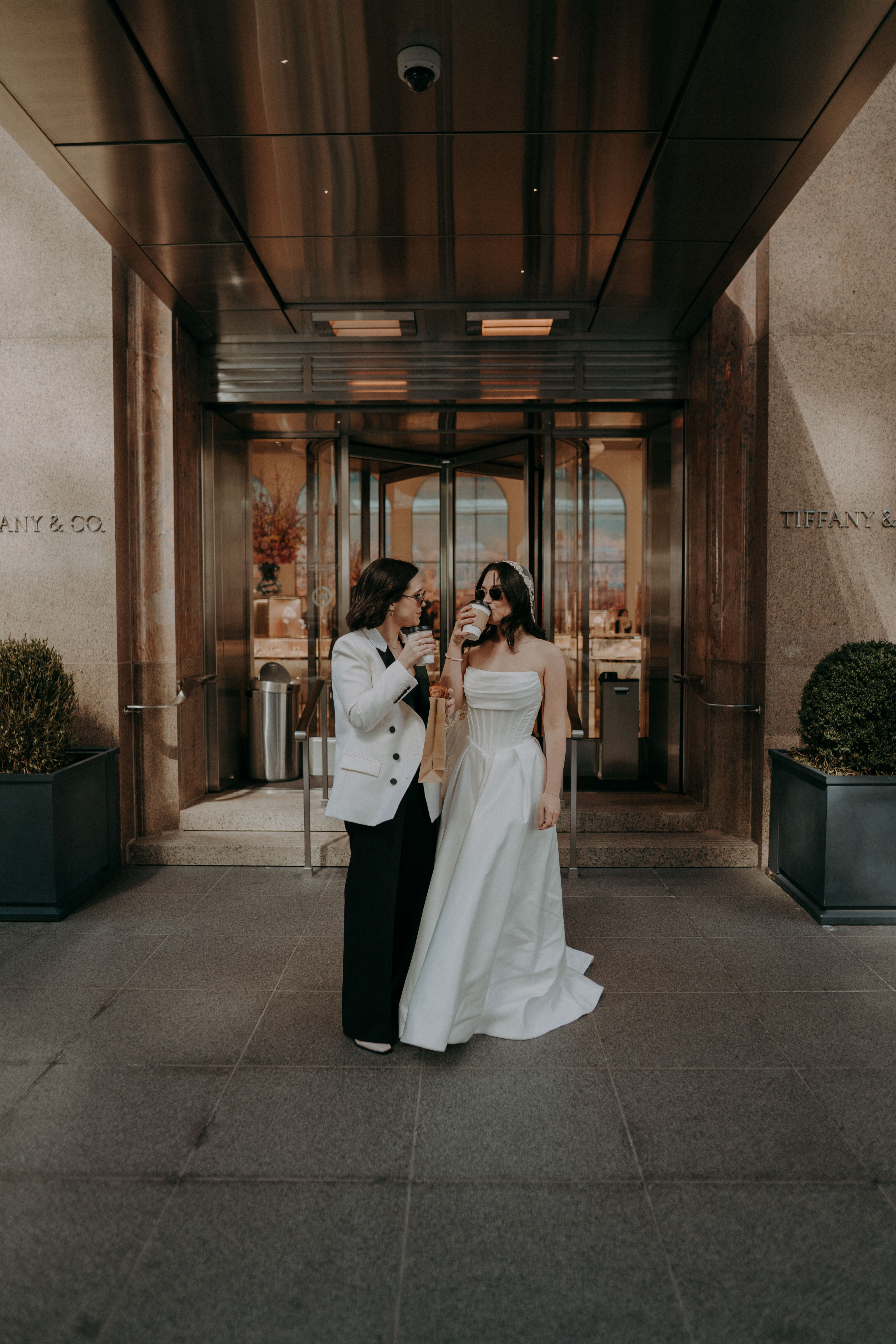 Two women dressed in wedding attire drinking coffee outside Tiffany & Co., a nod to Breakfast at Tiffany’s