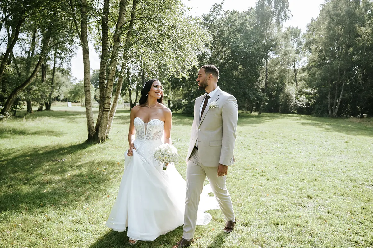 Brautpaar spaziert durch sonnigen Birkenpark, sie hebt das Kleid und lächelt ihm zu. Pärchenshooting hochzeit in natürlicher hochzeitsfotografie mit modernen hochzeitsfotos