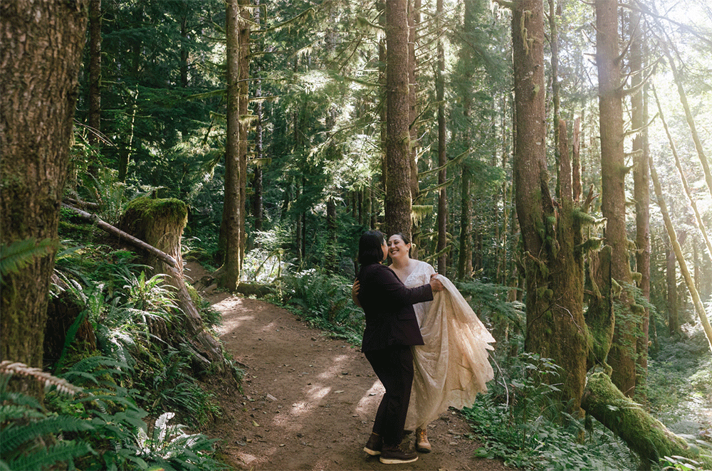 A Puerto Rican bride wearing a white gown with the sleeves off shoulder stands under a clear umbrella held by her groom in a black suit standing in front of Oregon's Multnomah Falls as the snow begins to fall for their winter adventure elopement. | Erica Swantek Photography