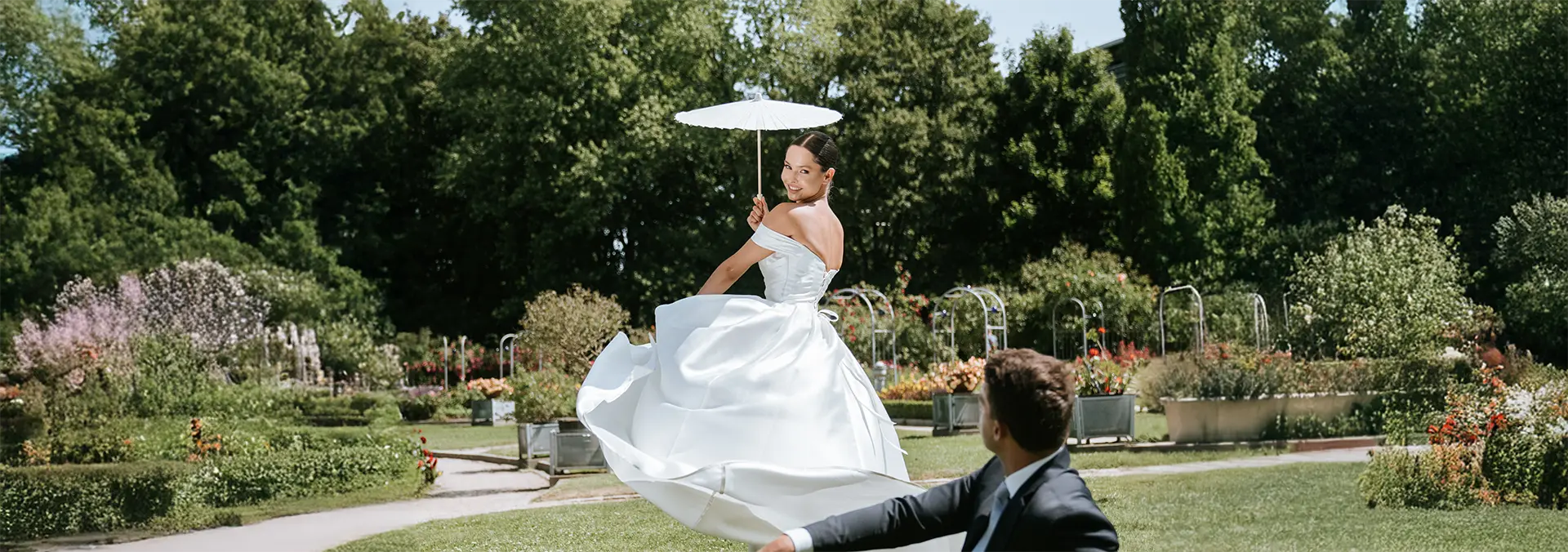 Die Braut wirbelt mit weißem Parasol durch die Parkwege, der Bräutigam schaut lächelnd zu; moderne hochzeitsfotografie und hochzeitsbilder draußen im Sommerlicht