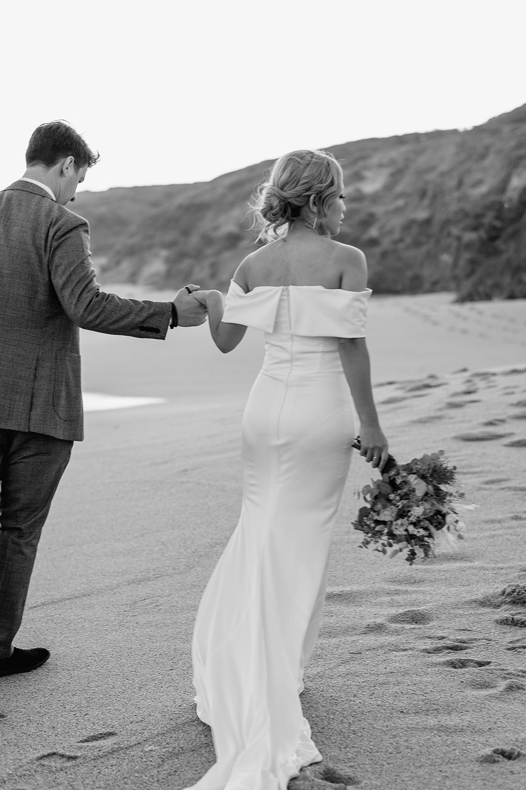 black and white photo of a woman and a man walking along the beach, holding flowers, about to get married