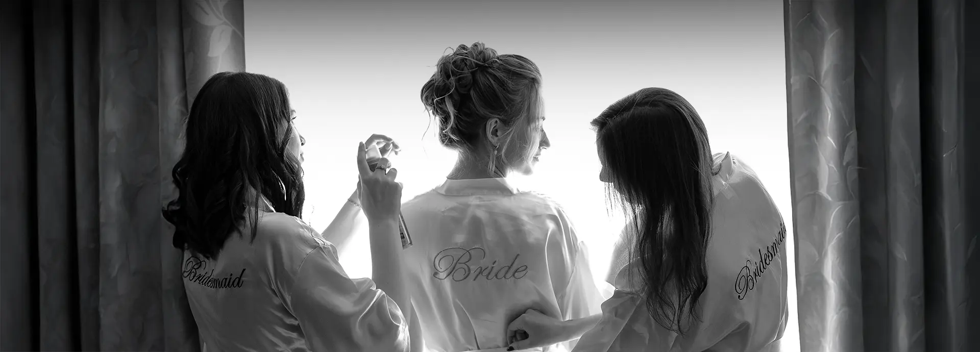 Braut im Morgenmantel am Fenster, zwei Brautjungfern helfen und sprühen Parfum; intimer Moment der getting ready fotografie, natürliche hochzeitsfotografie und hochzeitsfotograf mit stil in Köln