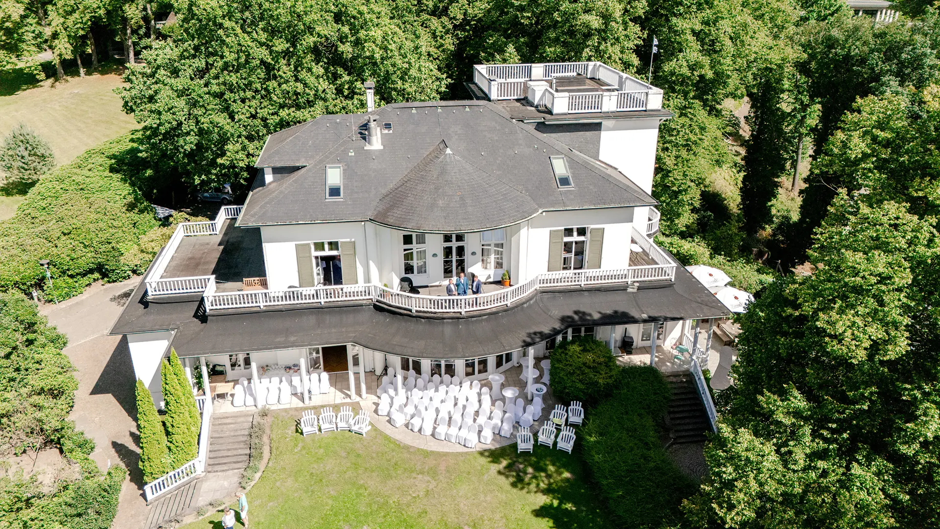 Fotograf mit Drohne – Luftaufnahme einer weißen Villa als Hochzeitslocation mit Terrasse und Garten.