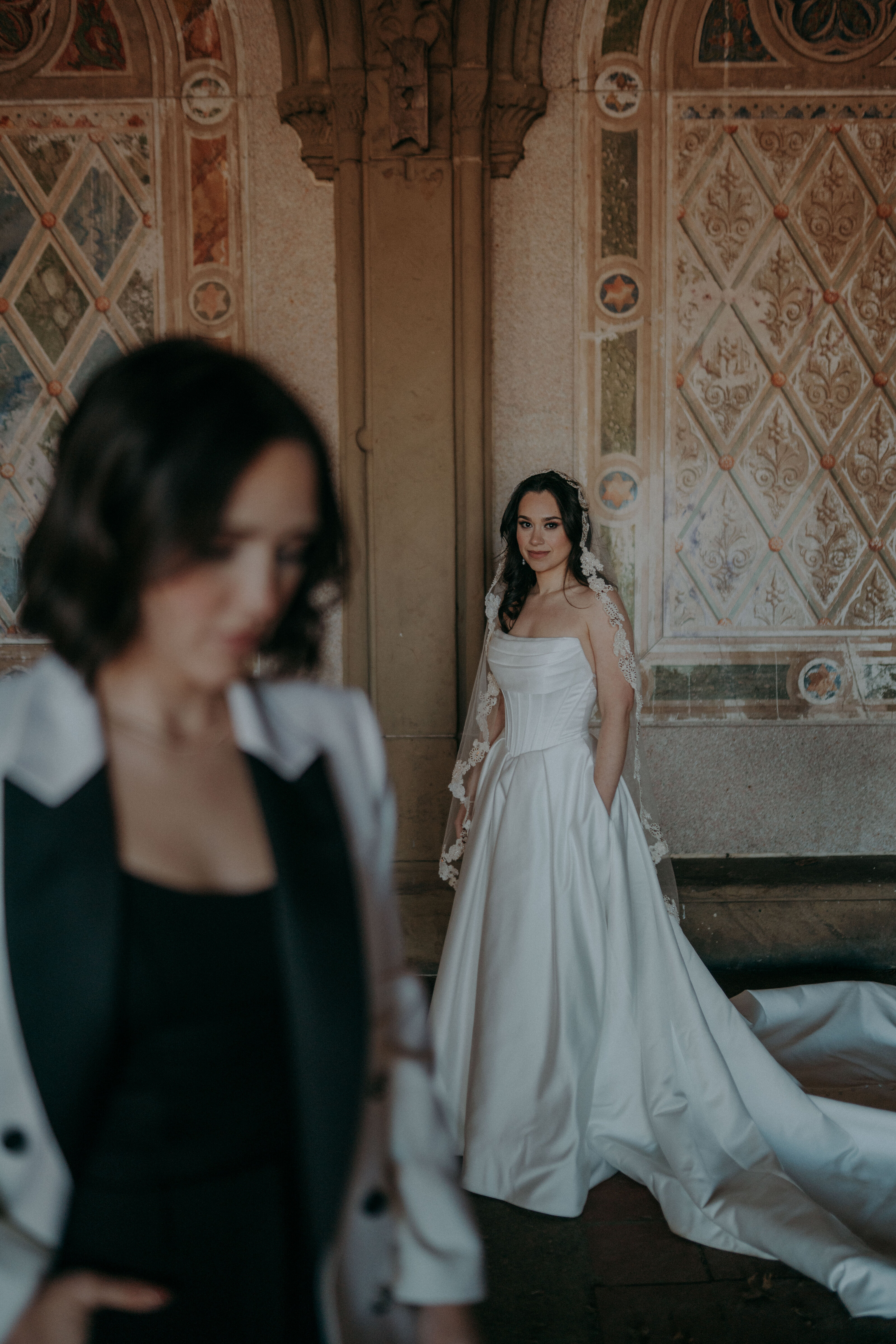A bride in a white gown stands against a decorated stone wall, with another woman in a white suit jacket blurred in the foreground.