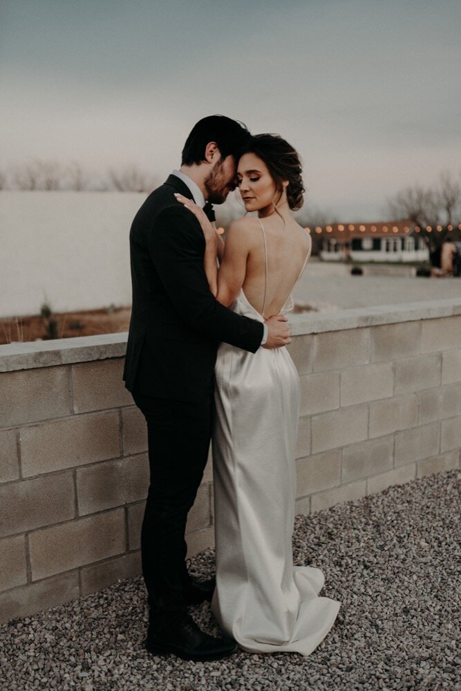 Couple embraces outdoors at sunset, bride in backless satin gown, groom in dark suit.
