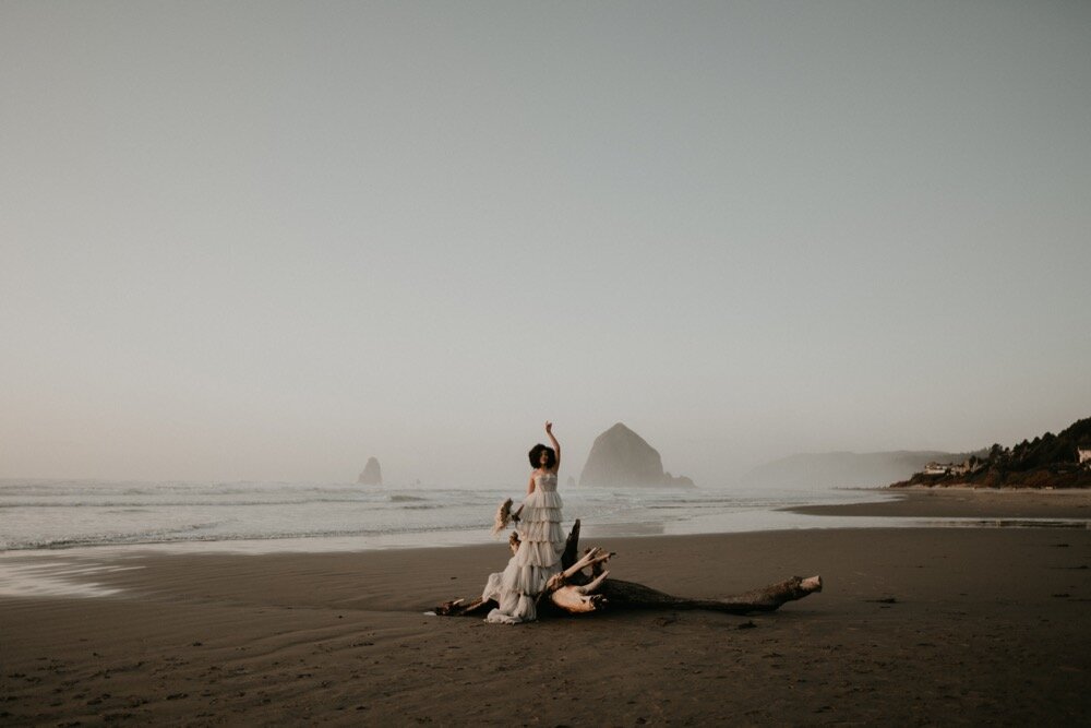 Bride in a ruffled gown standing on driftwood at a quiet beach with Haystack Rock in the distance, raising an arm gracefully into the air.