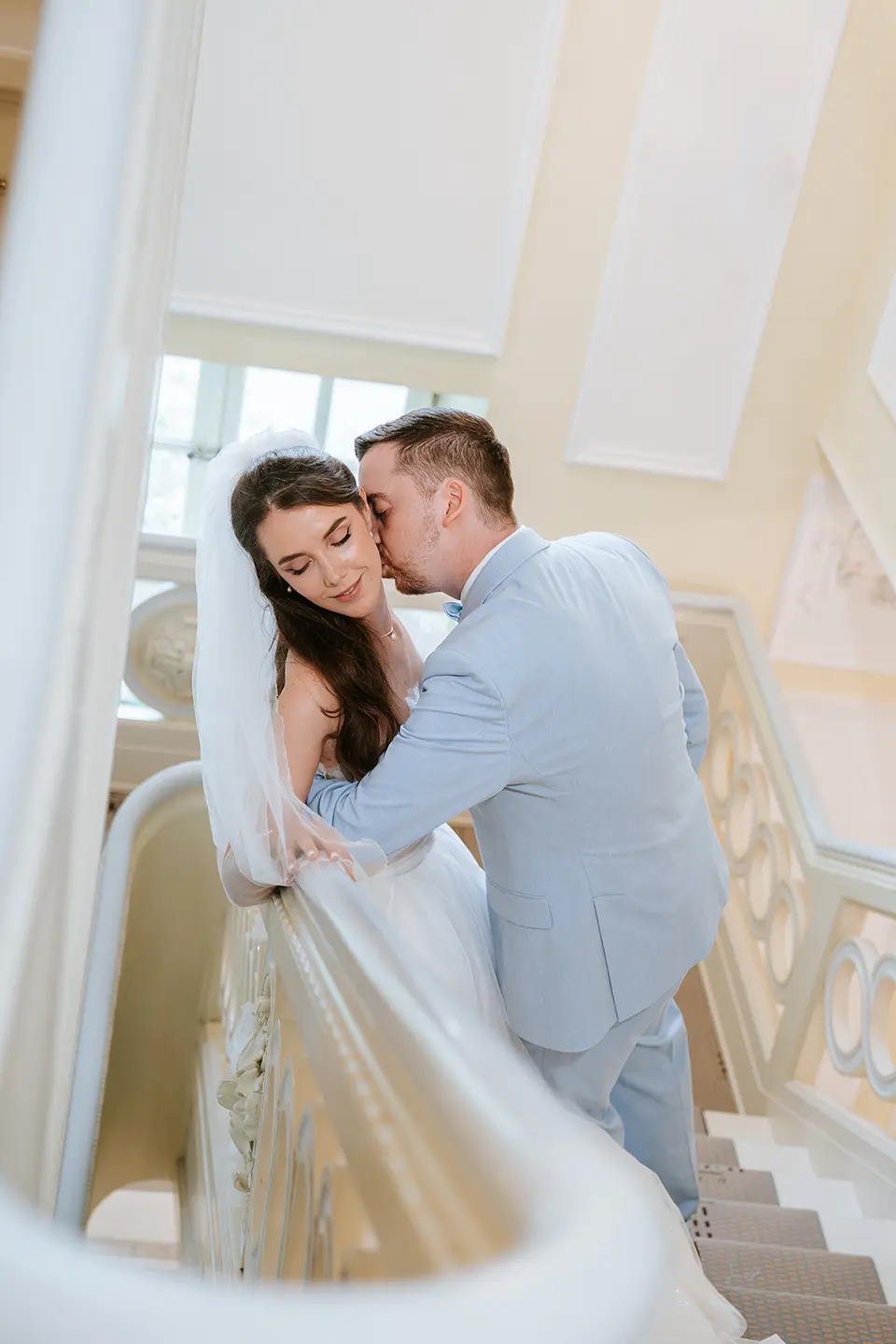 Braut und Bräutigam an einer hellen Treppe, sanfter Kuss an der Wange, Schleier am Geländer; kreative hochzeitsfotografie trifft auf emotionale hochzeitsbilder