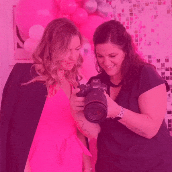 two  white women looking  into a camera. one is wearing a black shirt, one is wearing a pink shirt with a black jacket. The background is glittery with pink balloons.