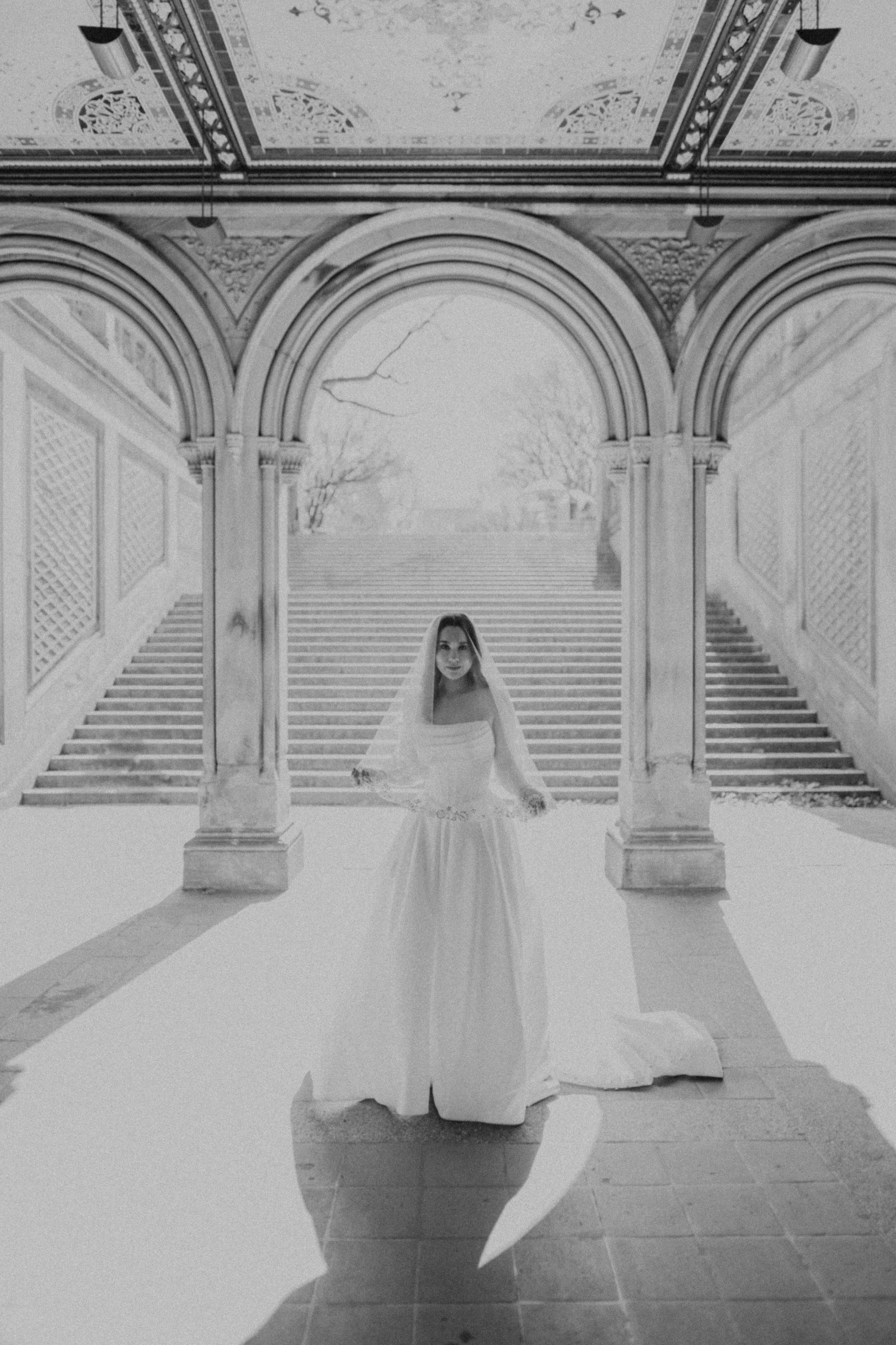Bride in lace-trimmed veil stands centered under ornate archway at Bethesda Terrace in Central Park.