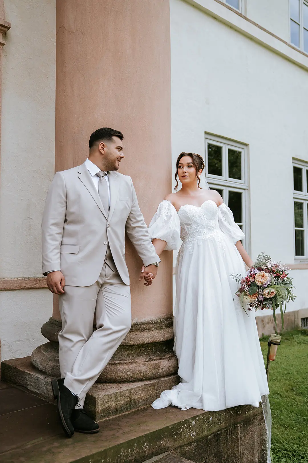 Das Paar hält Händchen an einer Sandsteinsäule, die Braut mit langer Schleppe und zartem Strauß – authentisch inszeniert als natürliche Hochzeitsfotografie mit Anklang an eine Schloss-Hochzeit