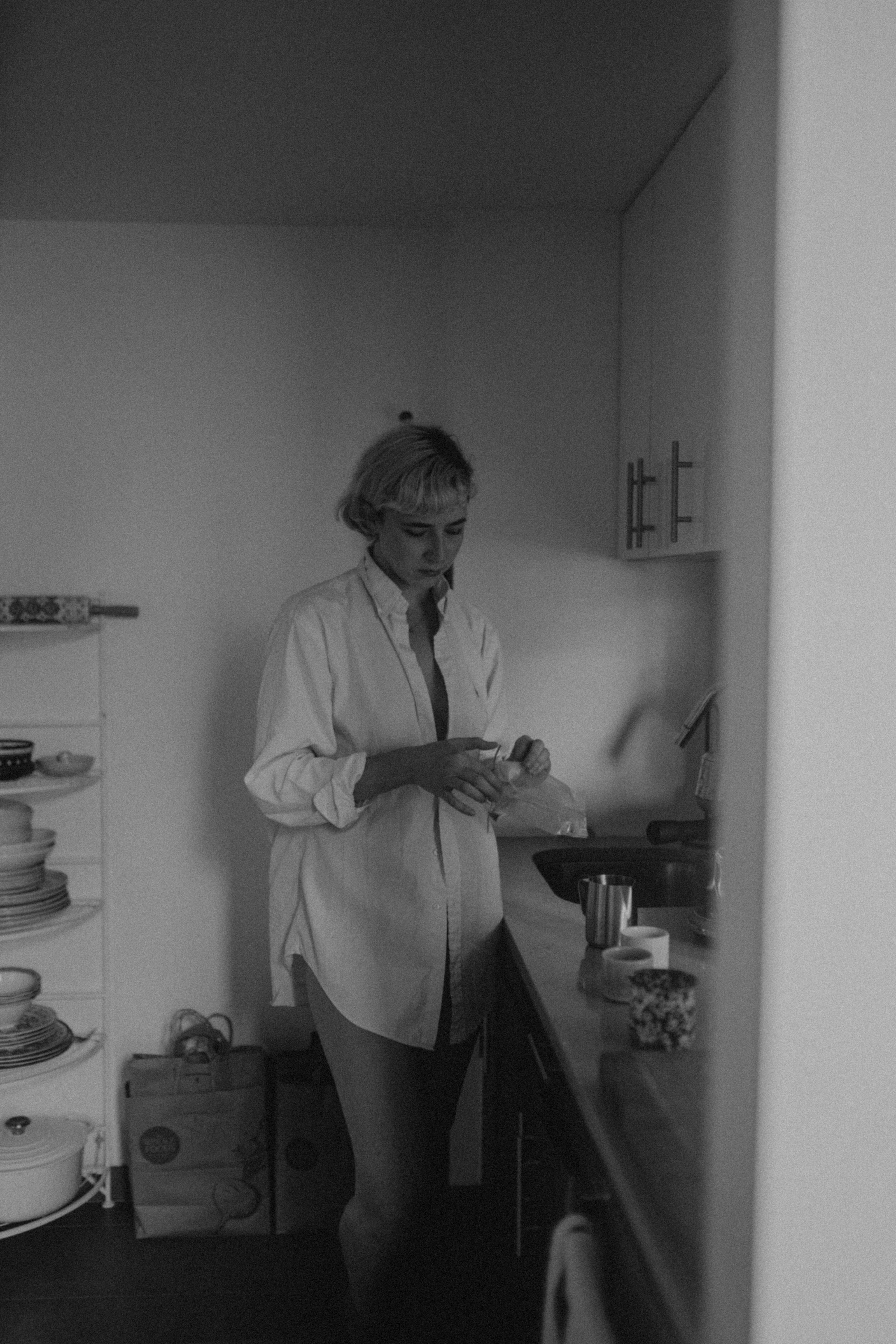 Woman with short blonde hair wearing an oversized white shirt, standing thoughtfully in a kitchen, holding a mug or glass.