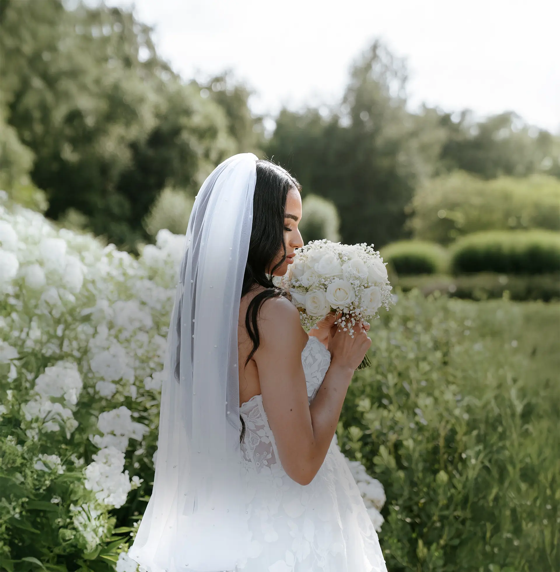 Braut im Profil mit langem Schleier, den weißen Rosenstrauß sanft am Gesicht; natürliche hochzeitsfotografie und fine art hochzeitsfotografie im blühenden Garten