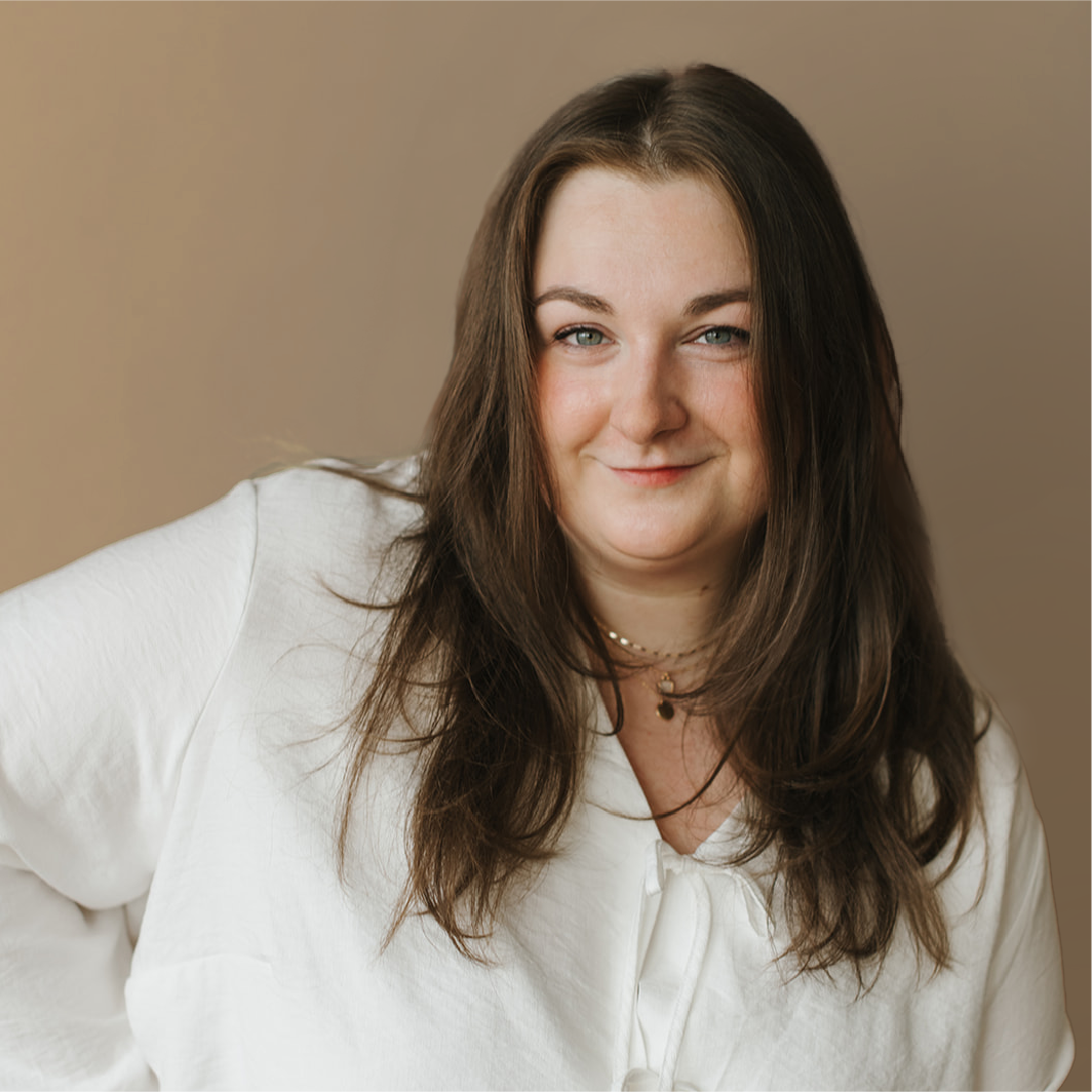 Woman with long brown hair wearing a white shirt, standing in front of a plain beige background and smiling slightly at the camera—perfect for a Showit designer or Showit design partner profile.