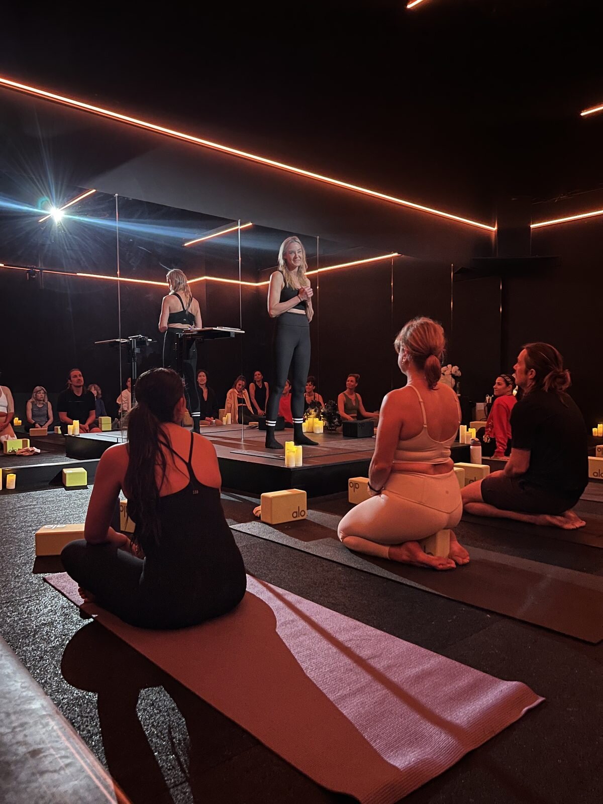Ceremny facilitator instructing from a candlelit platform under warm red LED lighting as seated students listen on yoga mats during an evening training session.