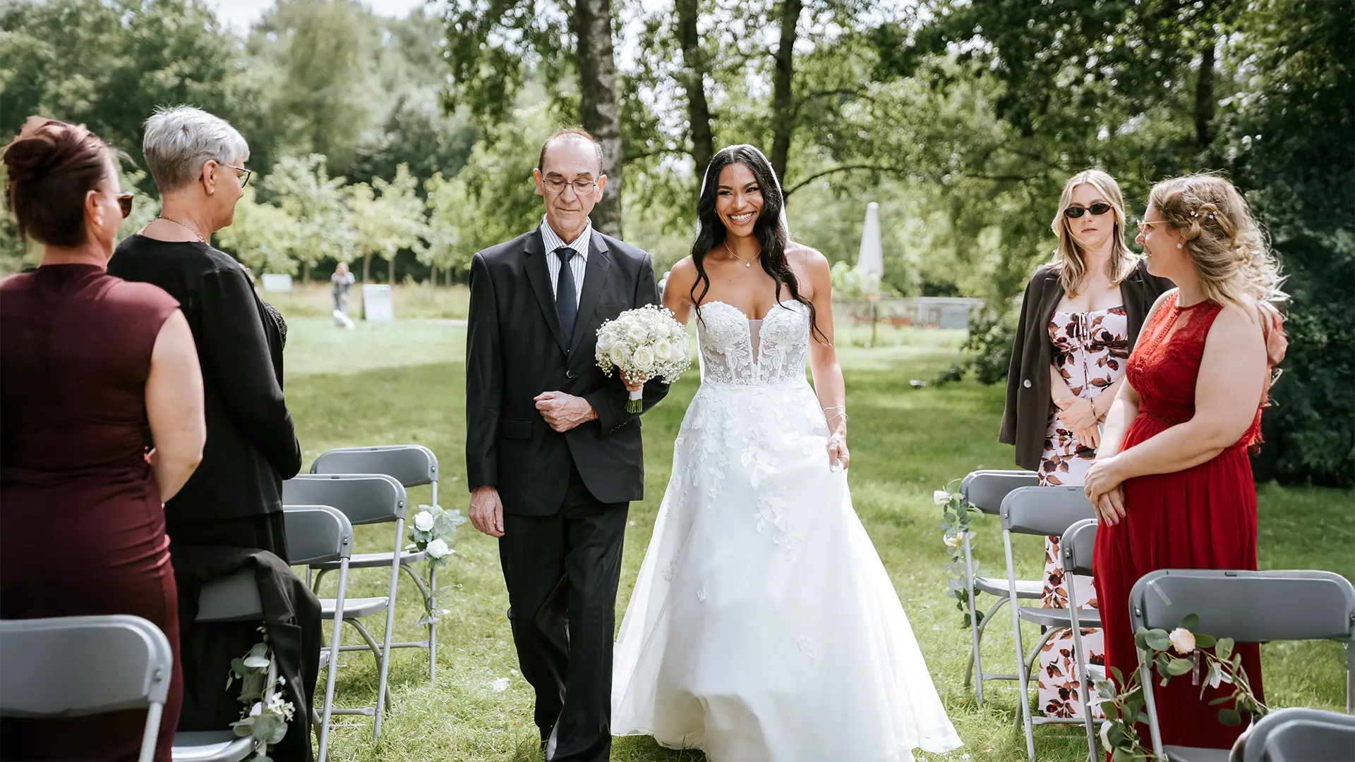 Die Braut schreitet mit Begleiter den Mittelgang entlang, Stuhlreihen flankieren den grünen Platz. Reportagefotografie Hochzeit und emotionale Hochzeitsbilder im Freien