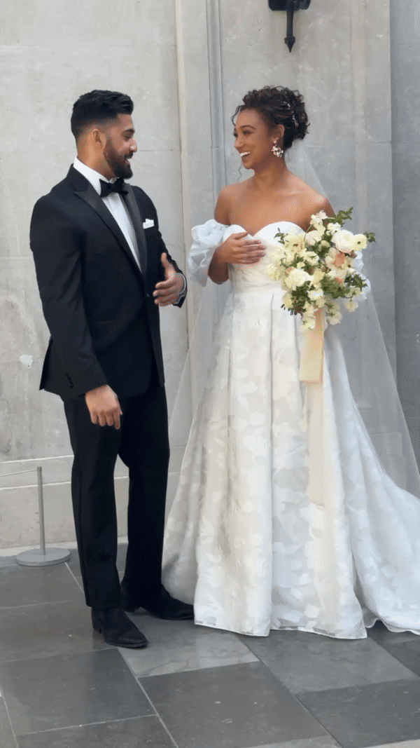 Bride in white gown smiles joyfully at groom in black tuxedo. She's holding a bouquet of white flowers, conveying happiness and celebration.