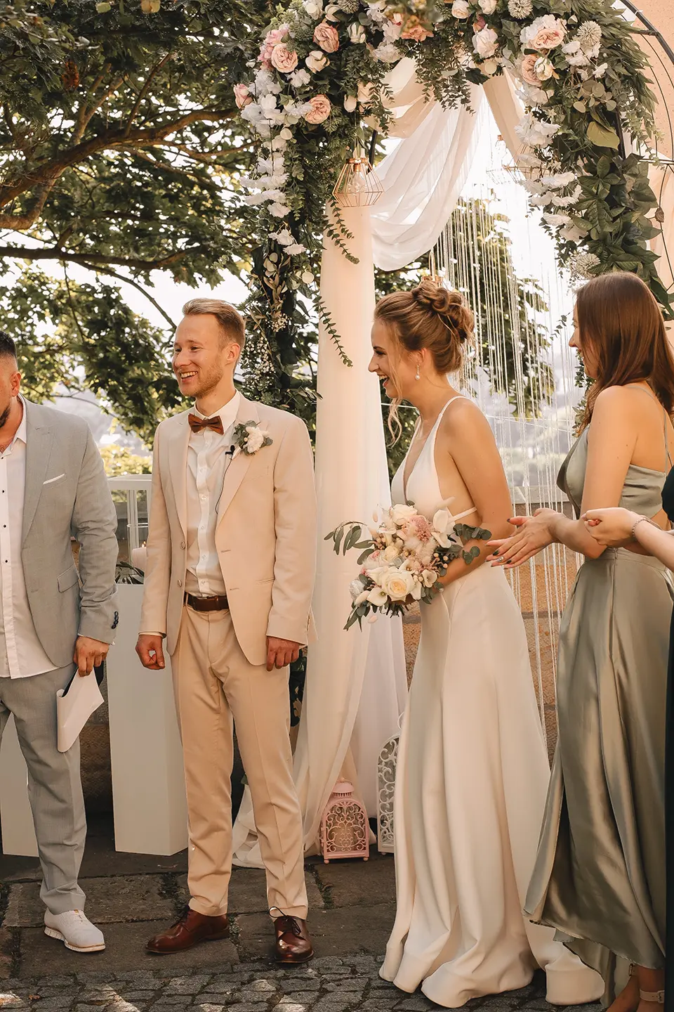 Braut und Bräutigam lachen unter einem üppigen Blumenbogen, Trauredner und Brautjungfer im Hintergrund; lebendige Szene für reportagefotografie hochzeit und hochzeitsfotografie mit stil, voller emotionaler hochzeitsbilder