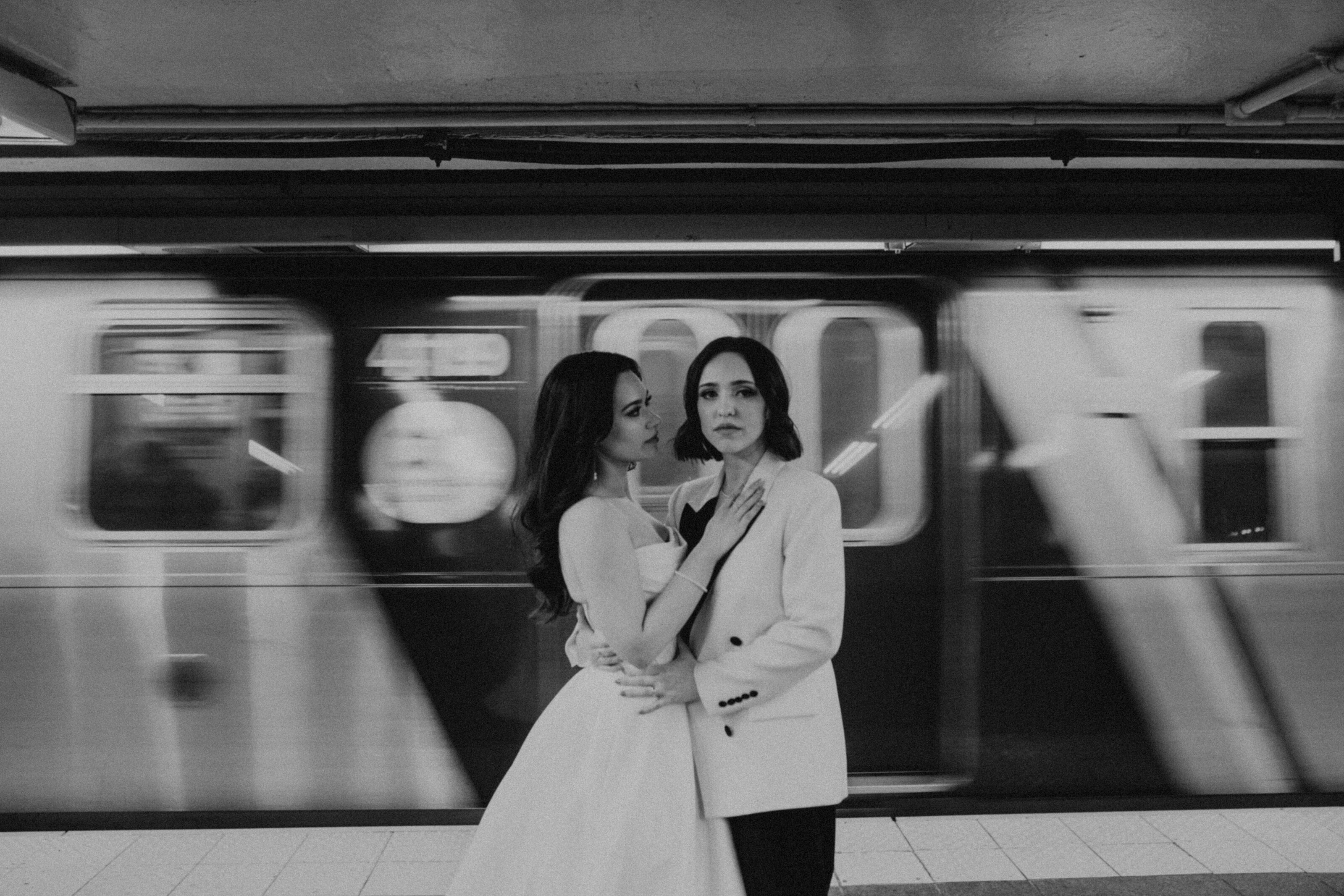 A bride and her partner stand on a subway platform, the train blurring behind them, the bride gazing at her partner.