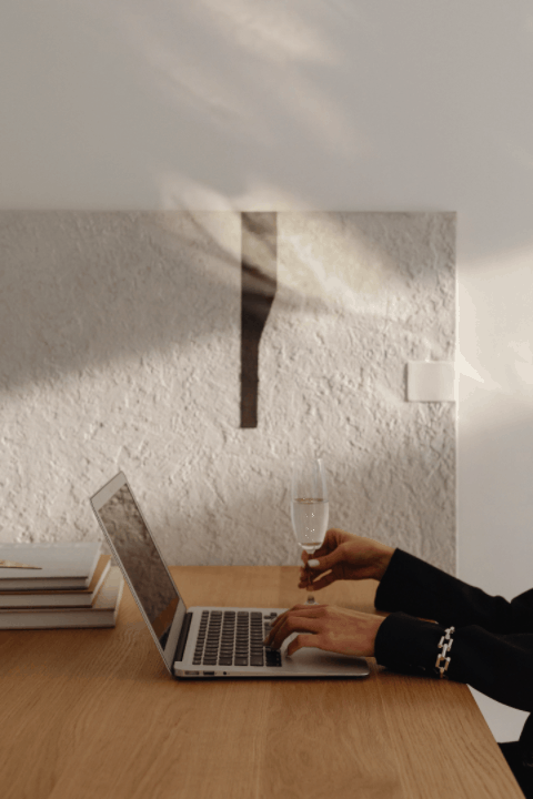 A stylish workspace featuring a laptop, stacked books, and a hand holding a champagne glass, evoking sophistication and refined productivity.