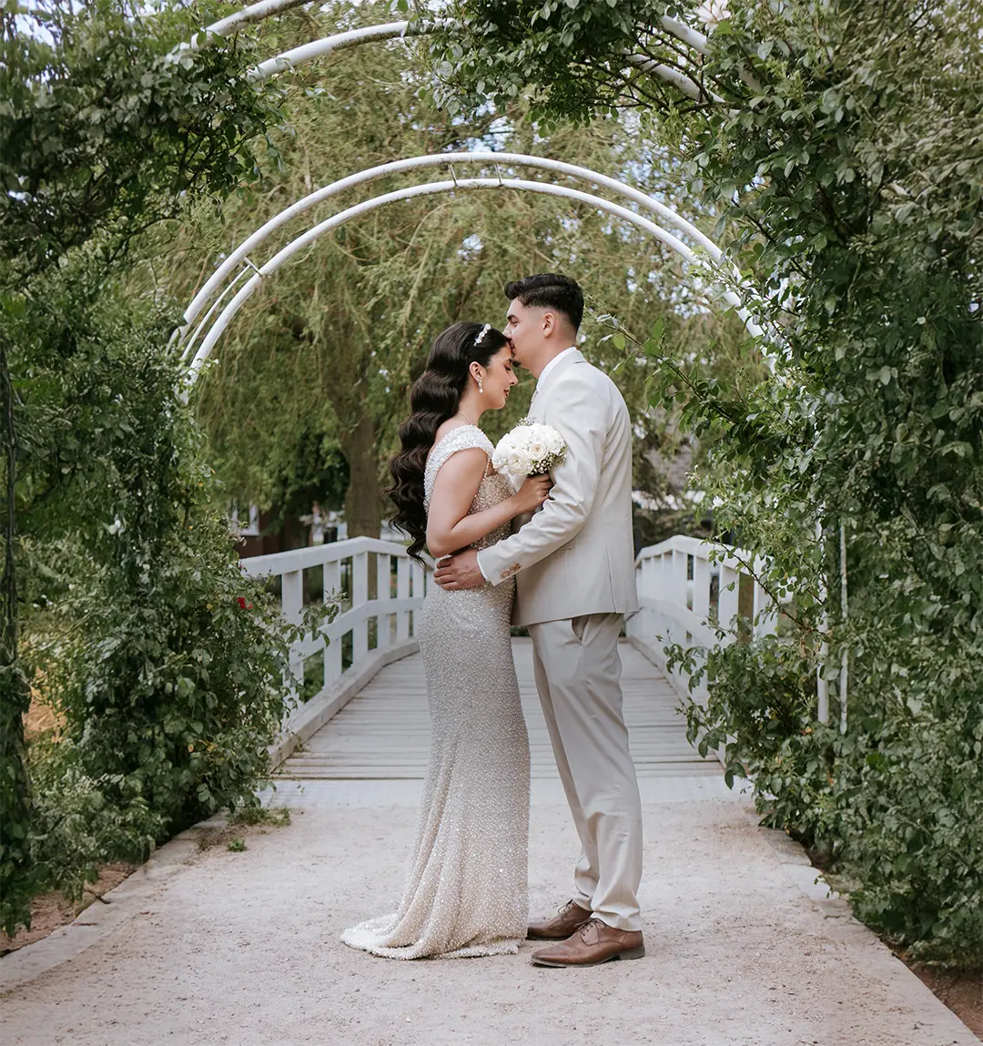 Zentral im grünen Laubengang stehen Braut und Bräutigam nah beisammen auf der Brücke, umgeben von Blättern und Licht; reportagefotografie hochzeit und moderne hochzeitsfotografie