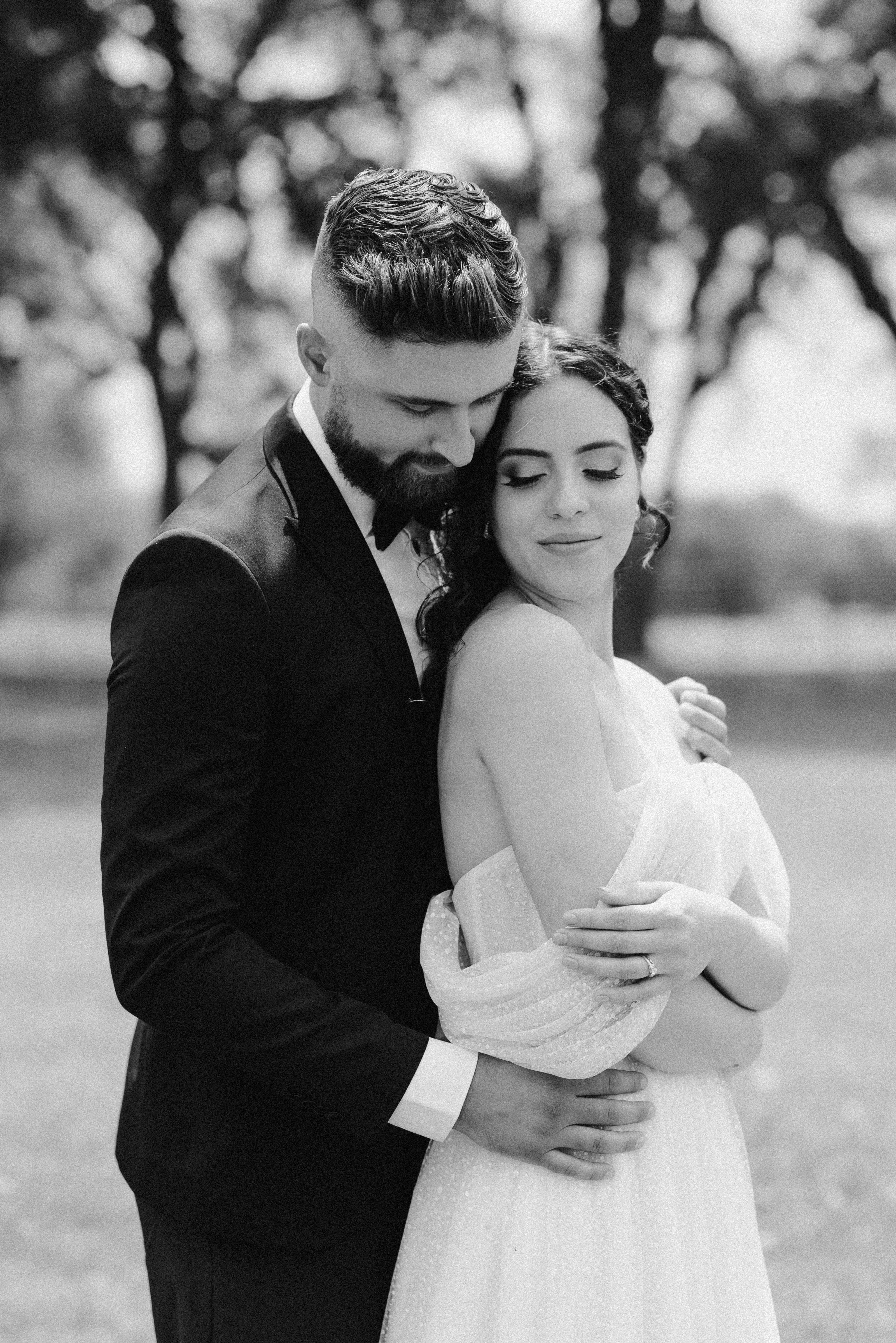 A black and white photograph captures a newlywed couple embracing in a serene outdoor setting. The image is a monochrome close-up shot of a man and a woman, presumably a newlywed couple, standing outdoors, embraced in a tender hug. 