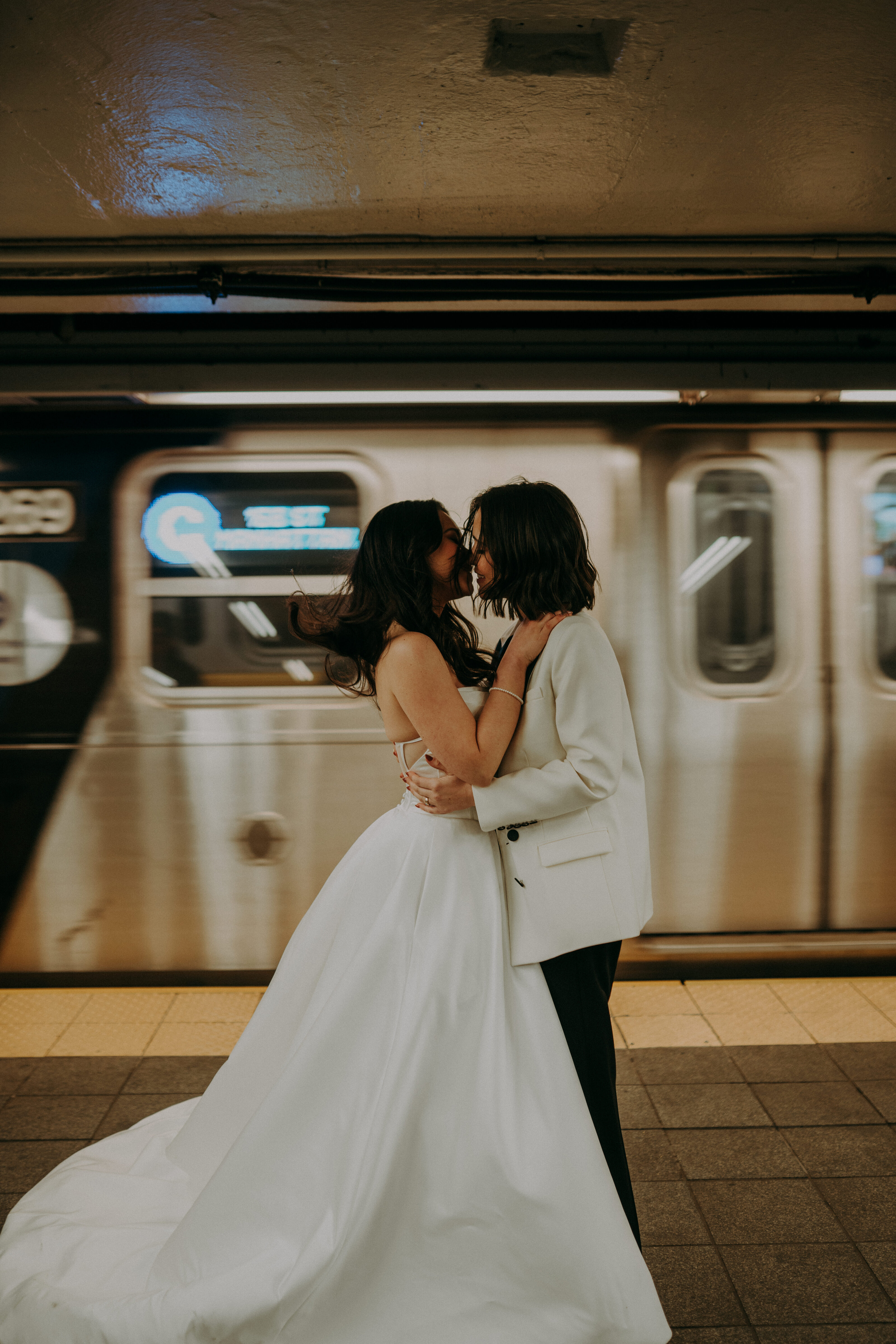 A couple in wedding attire kisses in a subway station as a train speeds by in the background.