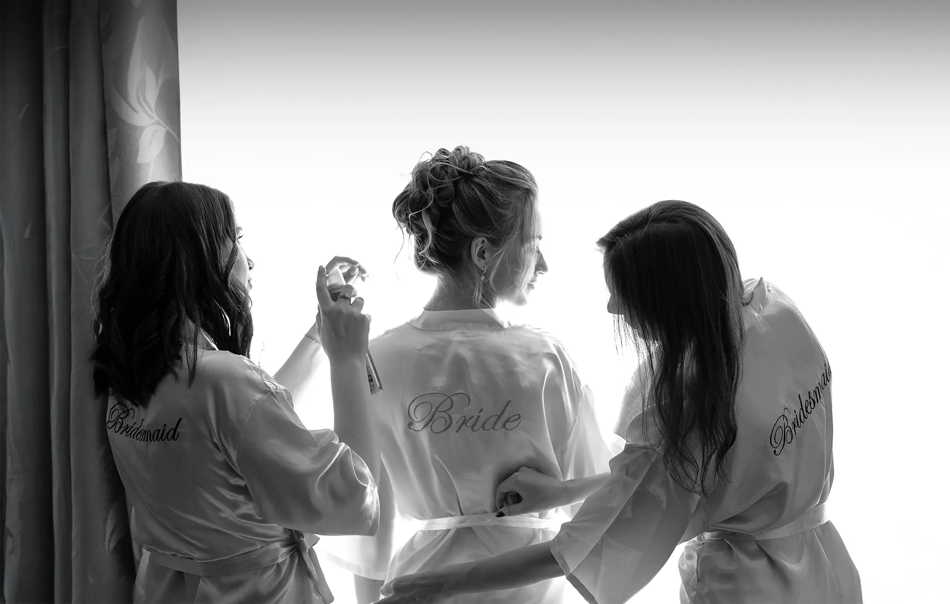 Braut im Morgenmantel am Fenster, zwei Brautjungfern helfen und sprühen Parfum; intimer Moment der getting ready fotografie, natürliche hochzeitsfotografie und hochzeitsfotograf mit stil in Köln