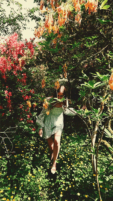 GIF of woman in pale blue dress walking outdoors under foliage before looking at the camera