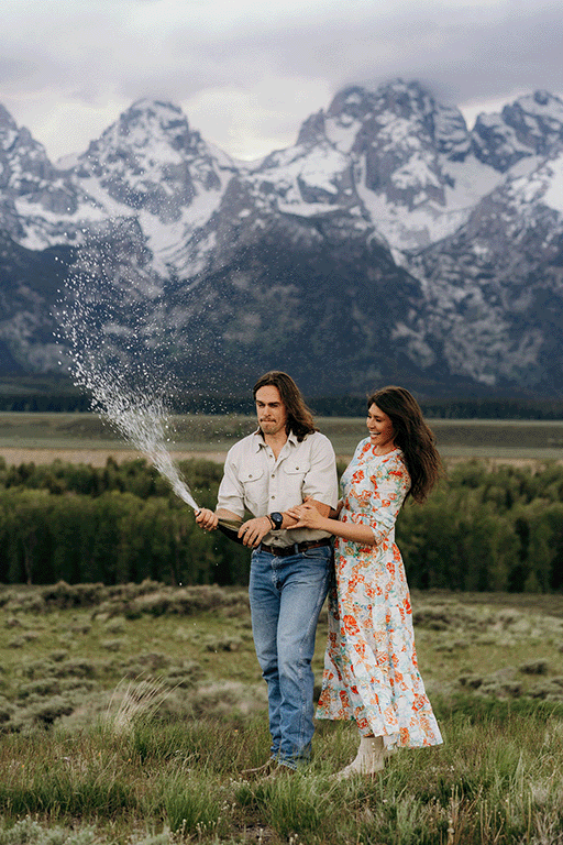 man and woman laughing while popping a bottle of champagne in a field with mountains in the background
