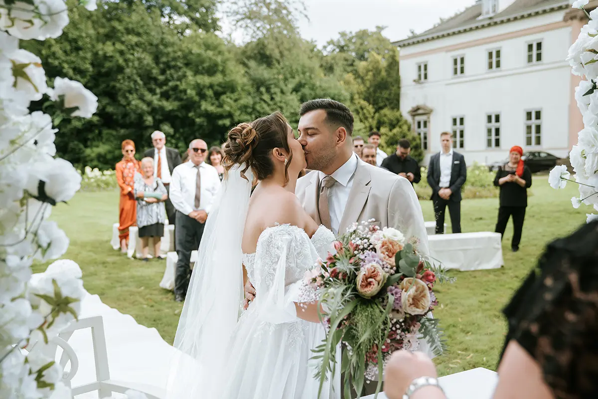 Während der freien Trauung im Garten küsst der Bräutigam seine Braut vor Familie und Freunden; künstlerische hochzeitsfotos und emotionale hochzeitsbilder Kiel