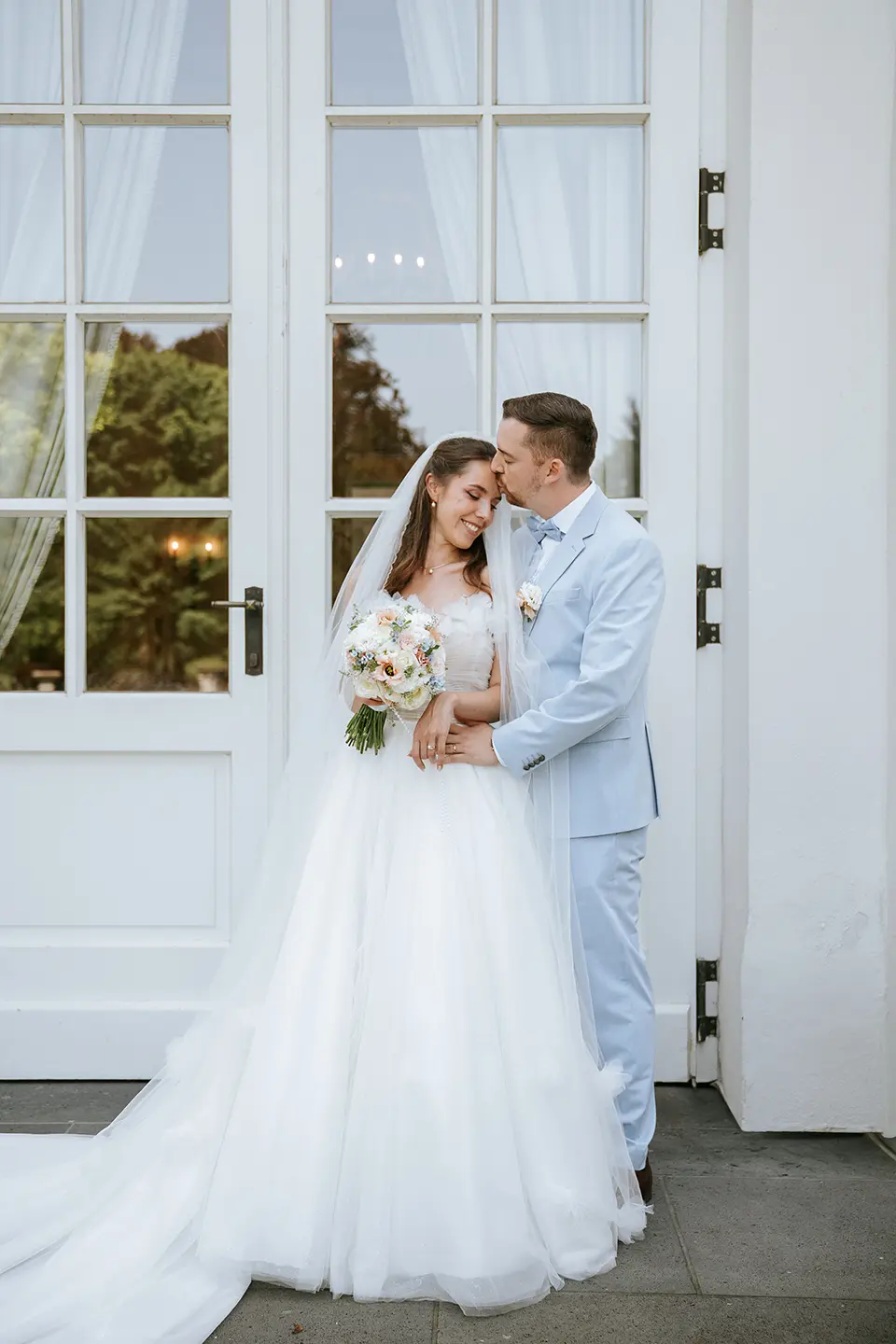 Brautpaar kuschelt vor weißen Flügeltüren, er küsst ihre Stirn, sie hält einen Pastellstrauß. Natürliche hochzeitsfotografie liefert emotionale hochzeitsbilder mit stil