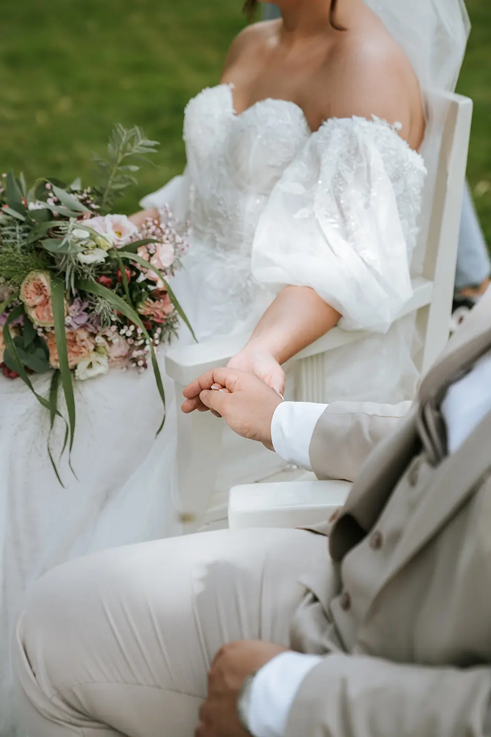 Detailaufnahme: Braut im Off-Shoulder-Kleid hält die Hand des Bräutigams, daneben ein üppiger Strauß im Garten; vertikales Mobil-Header-Bild – natürliche Hochzeitsfotografie, Hochzeitsbilder draußen, Hochzeitsfotografie mit Stil
