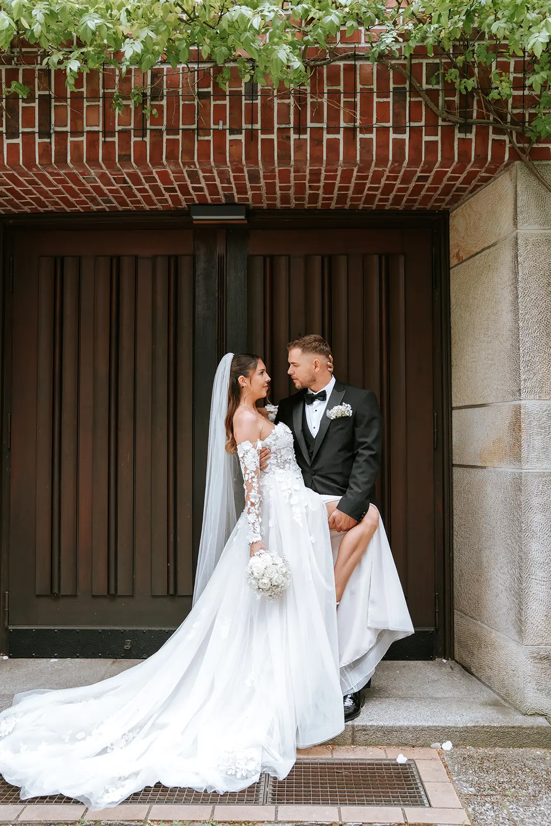 Vor dem dunklen Holztor posiert das Paar, die Braut trägt eine weite Schleppe und hält weiße Rosen; hochzeitsfotograf mit stil und künstlerische hochzeitsfotos