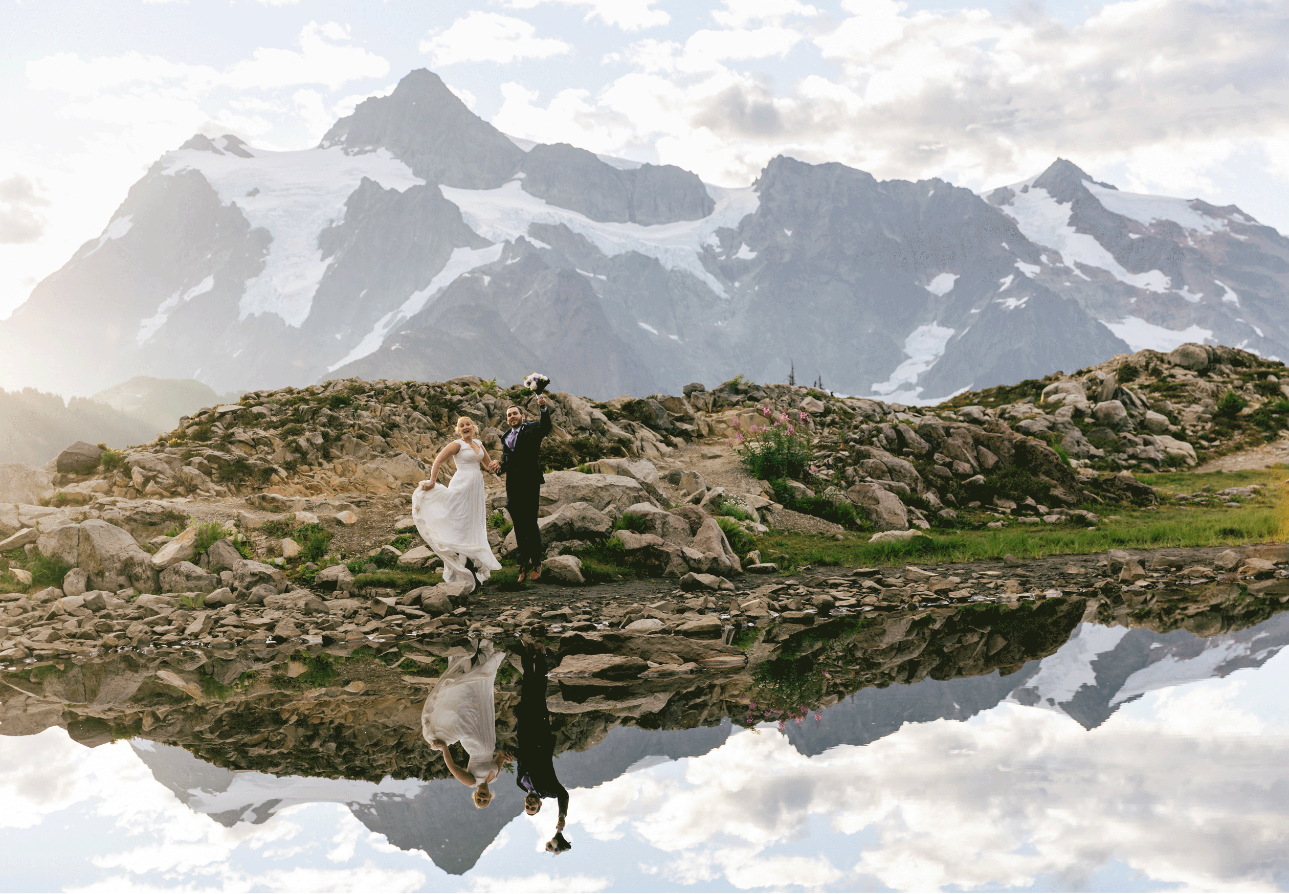 A blonde bride wearing a white flowing gown holds the skirt of her dress in one hand while holding her groom's hand in front of the reflection of Mt. Shuskan at Artist's Point in the North Cascades of Washington during their adventure elopement. | Erica Swantek Photography