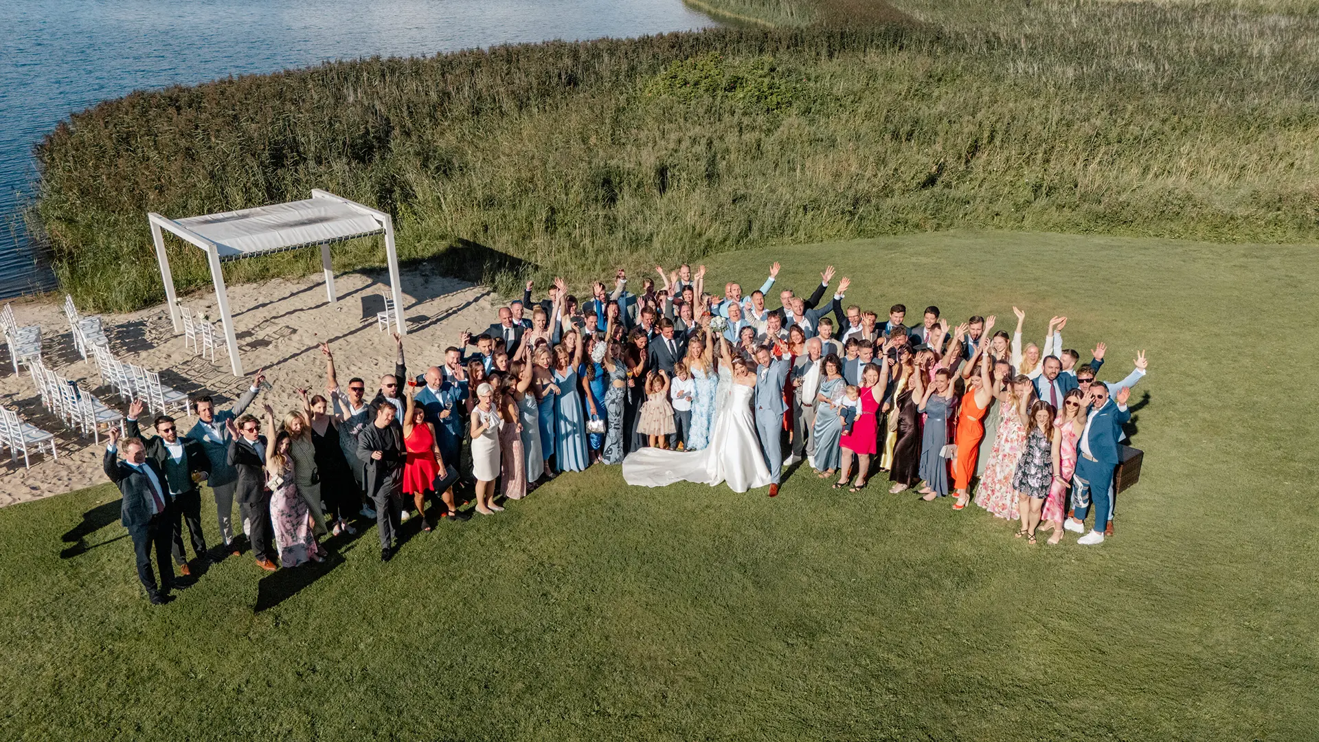 Drohnen-Fotograf – große Hochzeitsgesellschaft beim Gruppenfoto aus der Luft am Wasser und Strandpavillon.