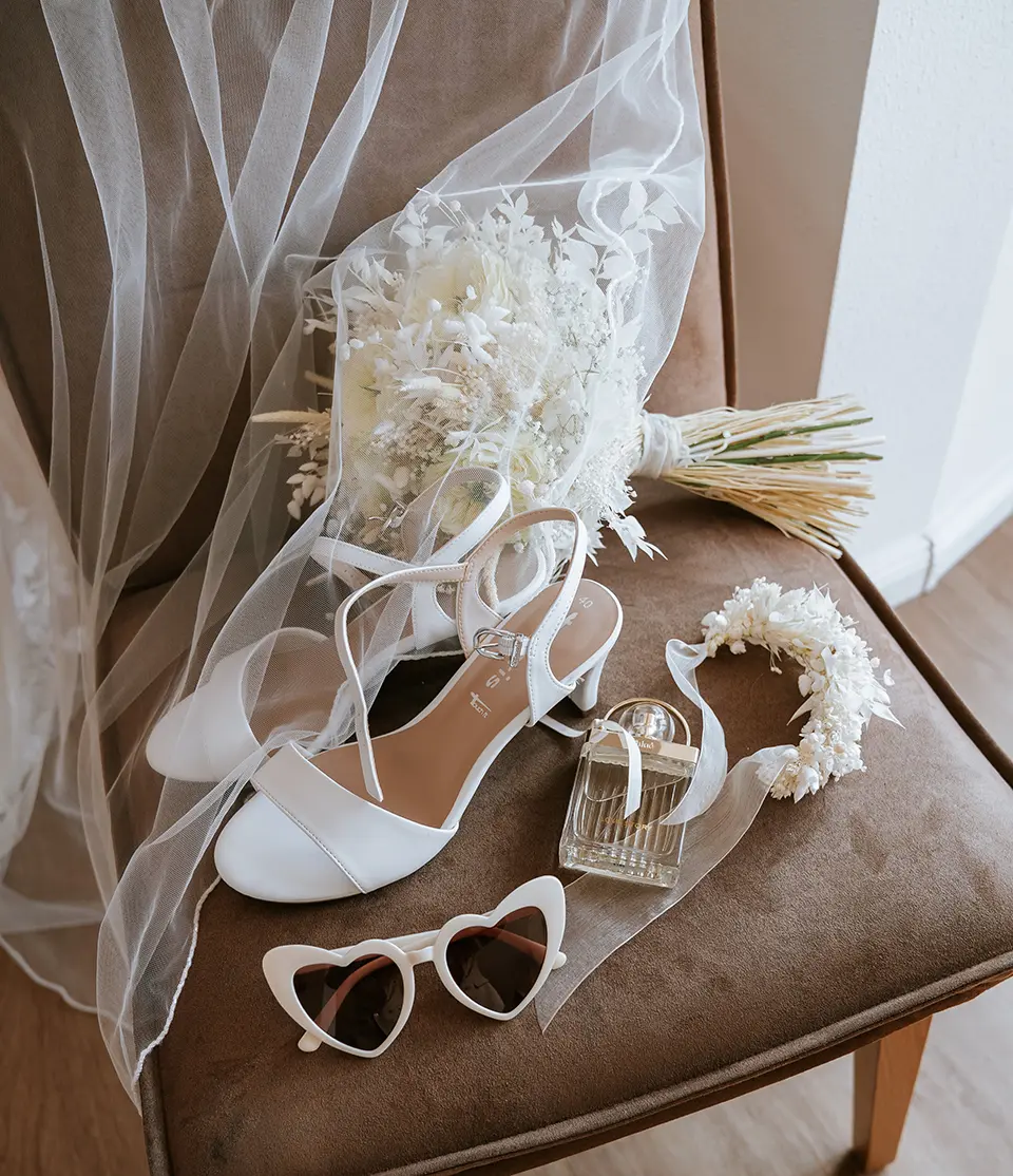 Weiße Heels, Schleier, trockener Strauß, Parfumflakon und Herzsonnenbrille harmonisch arrangiert. Getting ready fotografie kombiniert kreative hochzeitsfotografie mit hochzeitsfotograf mit stil