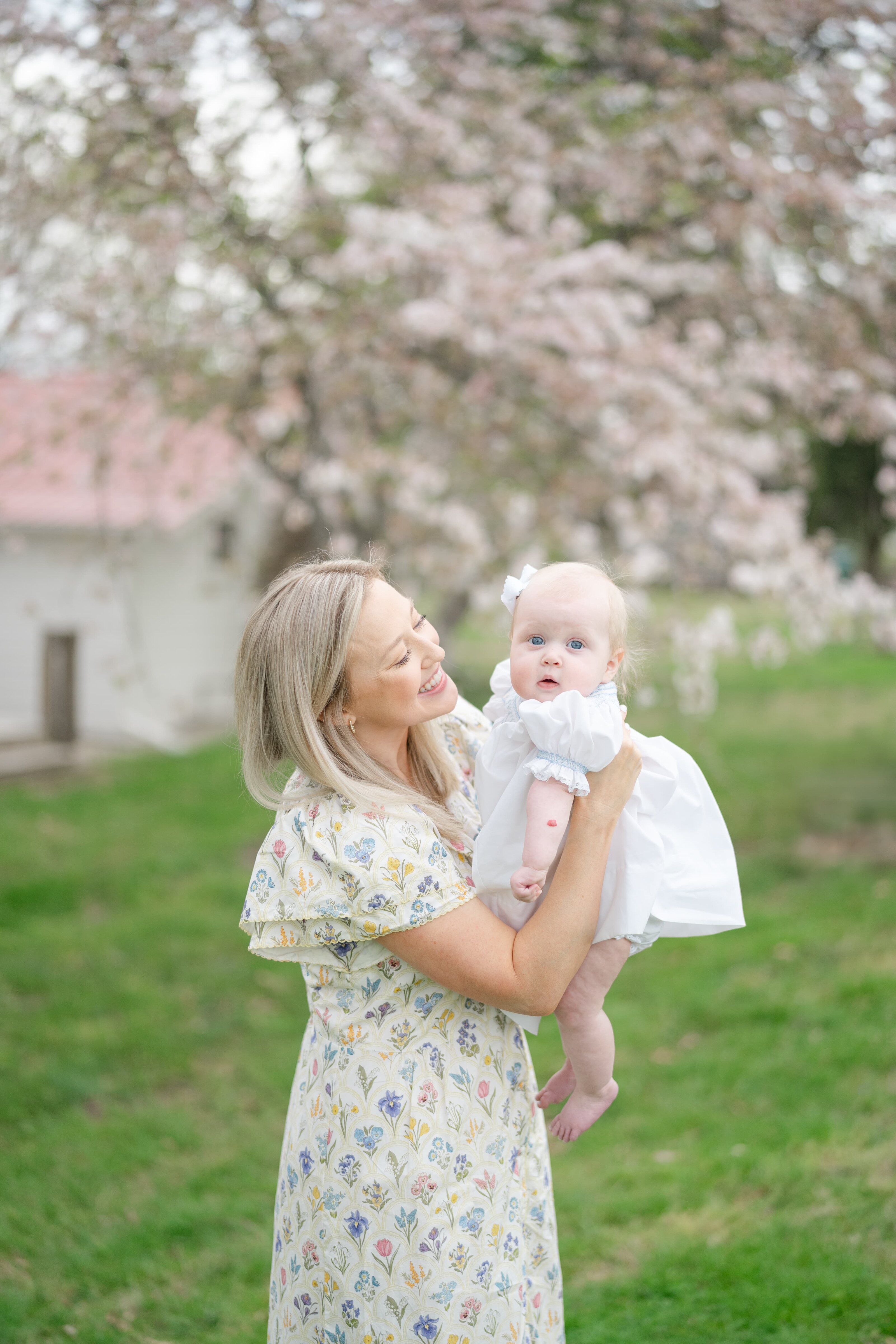Spring Family Photos At The Nashville Ellington Ag Center With Nashville Family Photographer Dolly DeLong A Mom Is Holding Her Baby Girl And Is Smiling With Spring Blossoms In Backdrop