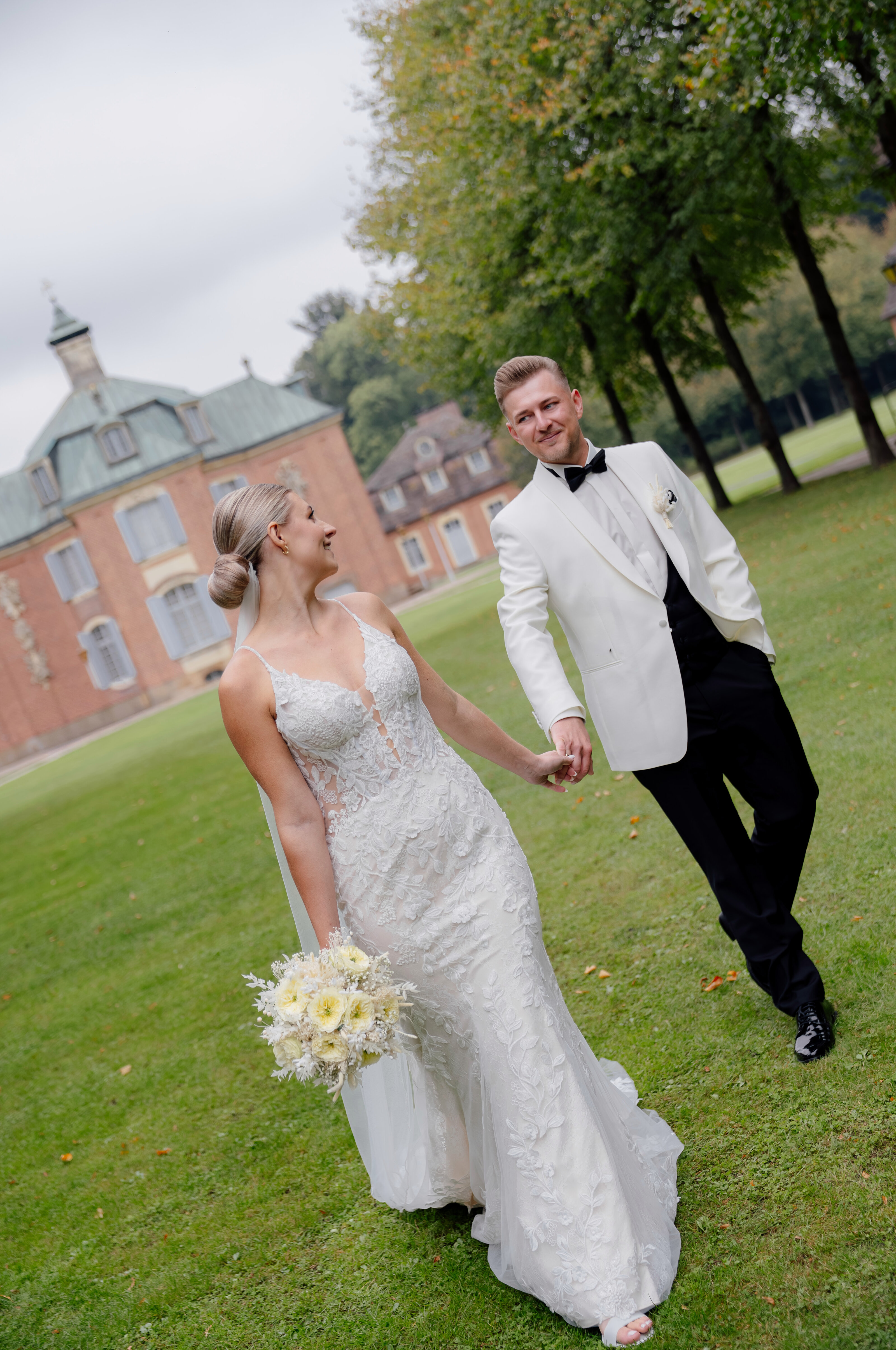 Braut und Bräutigam stehen sich liebevoll im Garten gegenüber; weißes Kleid und blauer Anzug inmitten grüner Bäume – romantische hochzeitsreportage lindern mit natürlicher hochzeitsfotografie