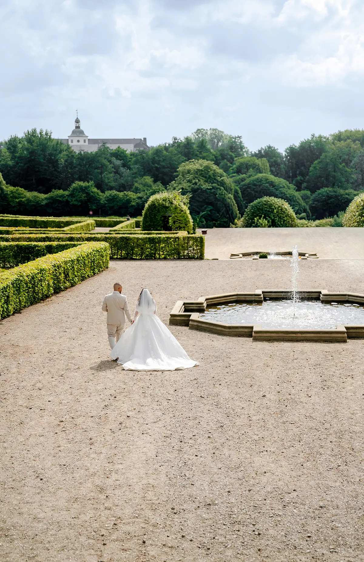 Drohnenaufnahmen Hochzeit – Brautpaar spaziert Hand in Hand durch eine geometrische Gartenanlage mit Brunnen.