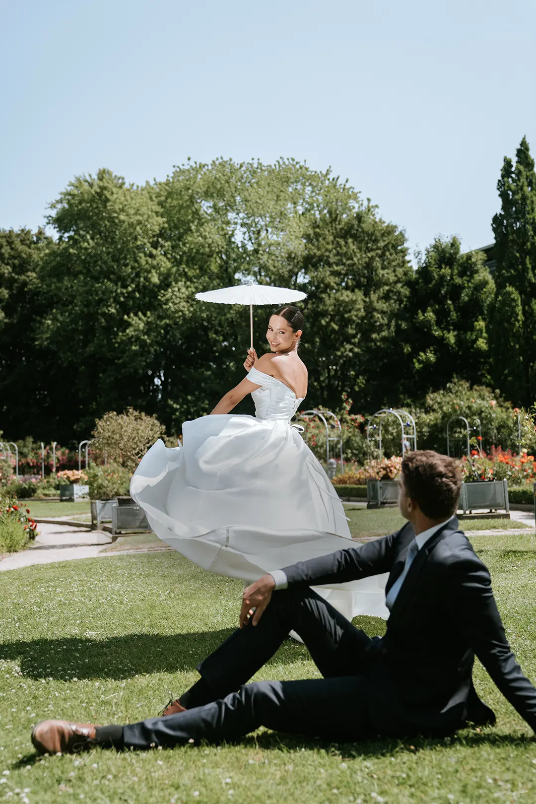 Vertikales Mobil-Header-Bild: Braut dreht sich mit weißem Schirm, Bräutigam sitzt im Garten; sommerliches Paarmoment in Düsseldorf – natürliche Hochzeitsfotografie, hochzeitsbilder draußen