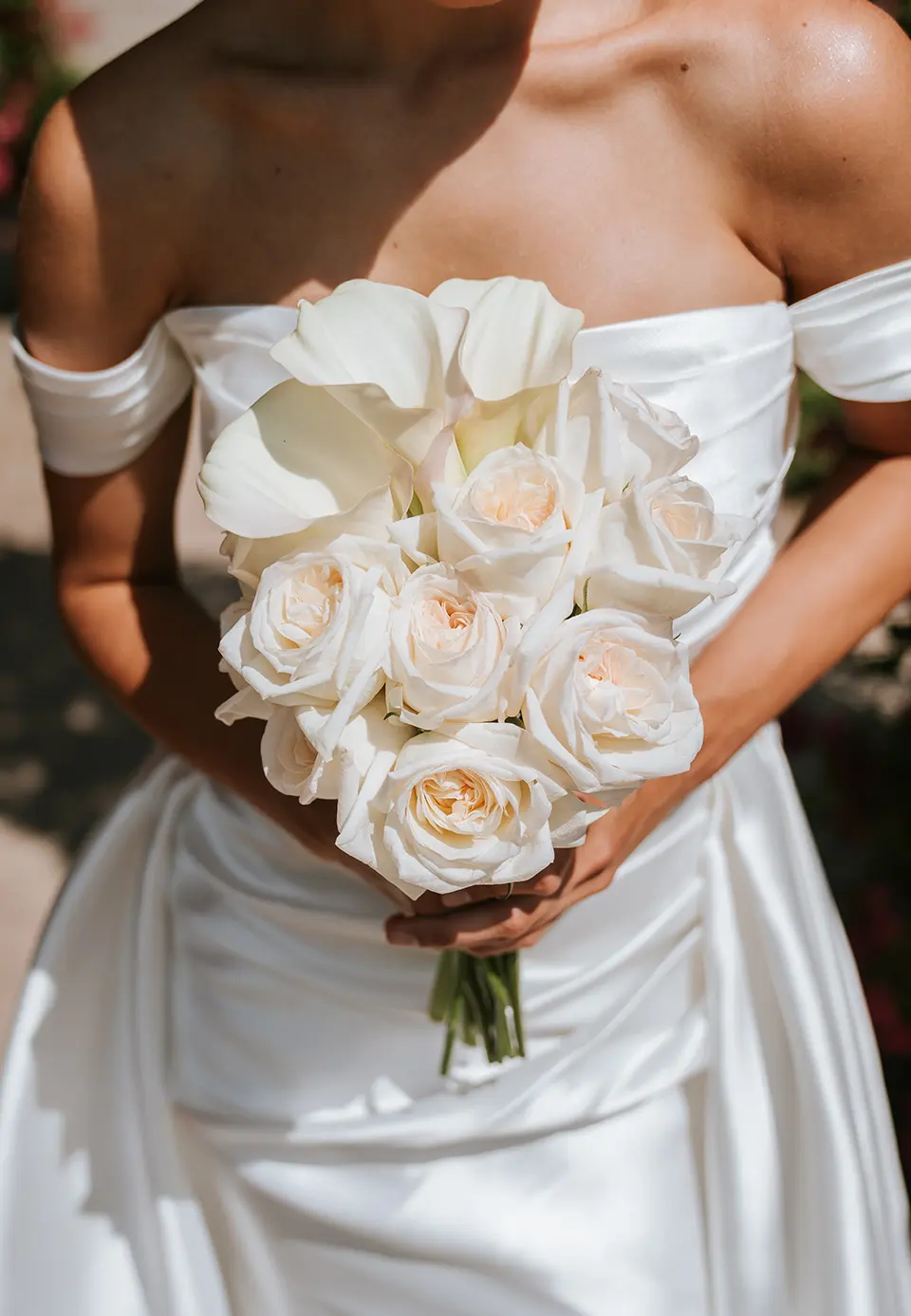 Zart gebundener Brautstrauß aus weißen Rosen und Calla, gehalten vor einem Off-Shoulder-Satinkleid; kreative hochzeitsfotografie und künstlerische hochzeitsfotos im Garten