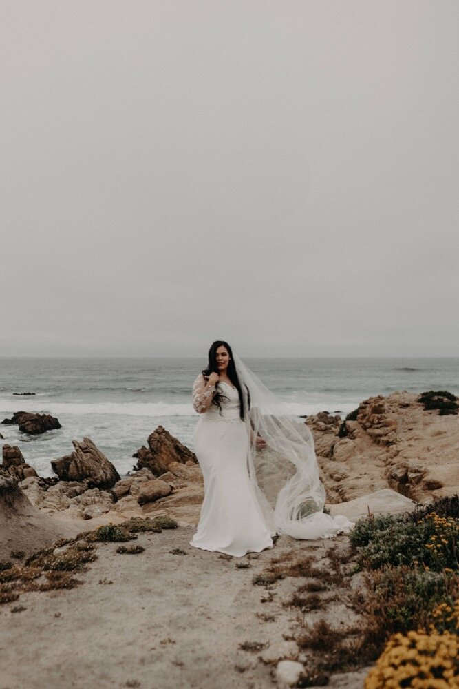 Bride standing on rocky cliff with waves and ocean in the background, long veil blowing in the wind.