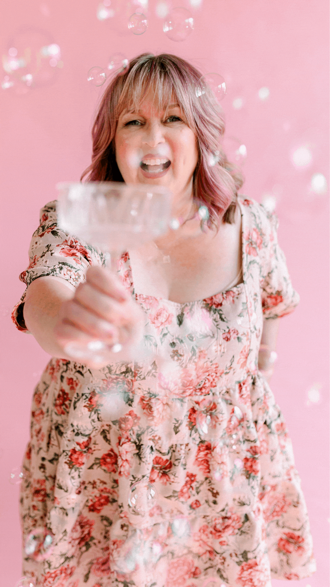 Woman with pink hair and white skin is sipping champagne and laughing in floral pink party dress, in front of a pink backdrop.