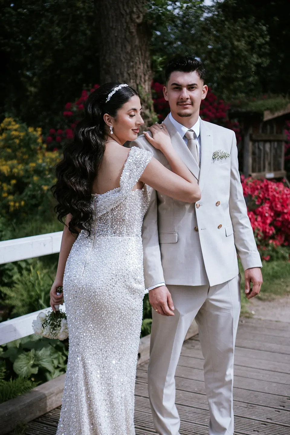Die Braut lehnt sich an den Bräutigam auf einer Holzbrücke, zeigt das perlenbesetzte Kleid und einen Blumenstrauß; kreative hochzeitsfotografie und klassische hochzeitsfotos