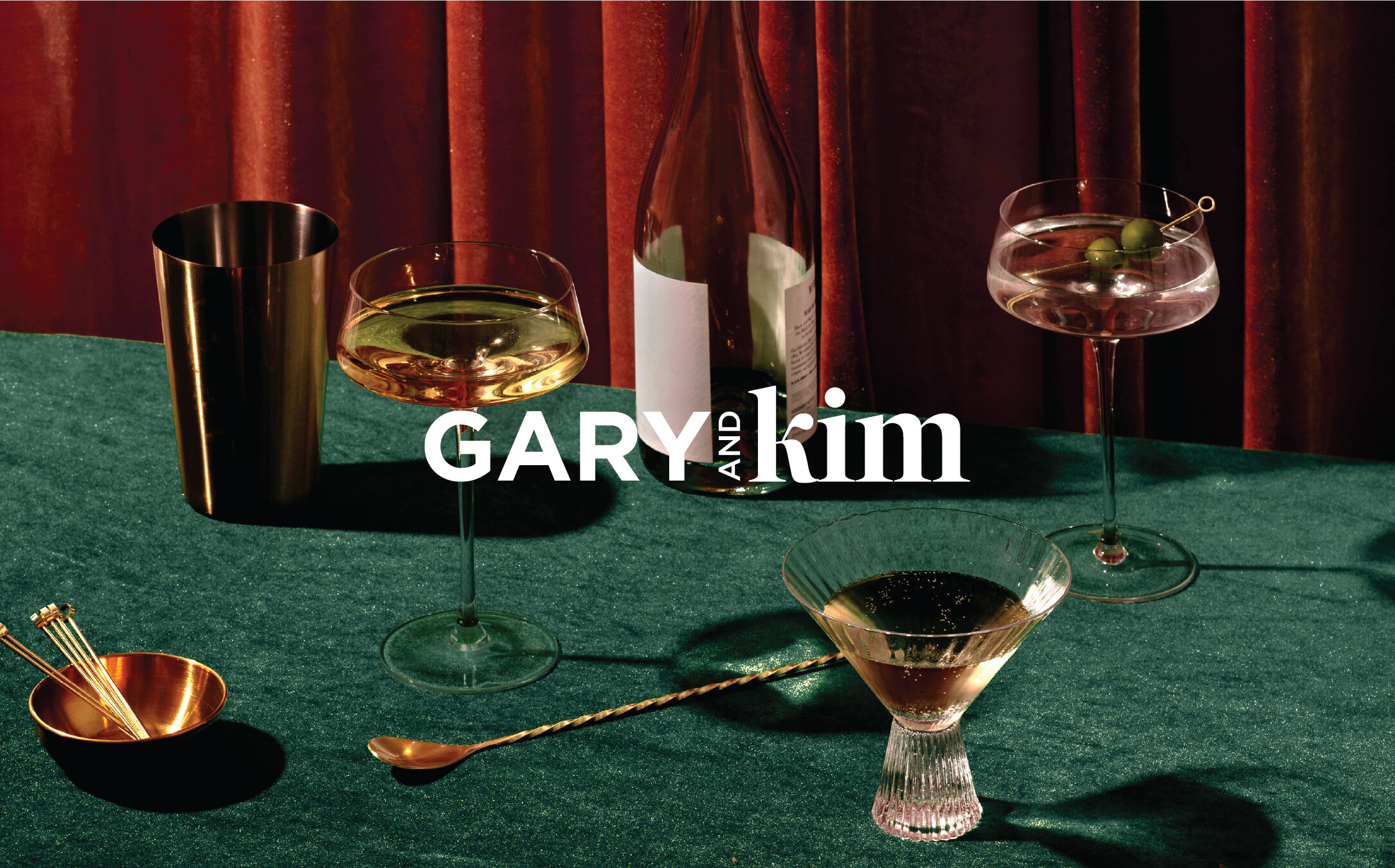 Primary logo for Gary and Kim overlaid on a moody cocktail scene with velvet tablecloth and vintage glassware, reflecting the brand’s upscale, immersive aesthetic.